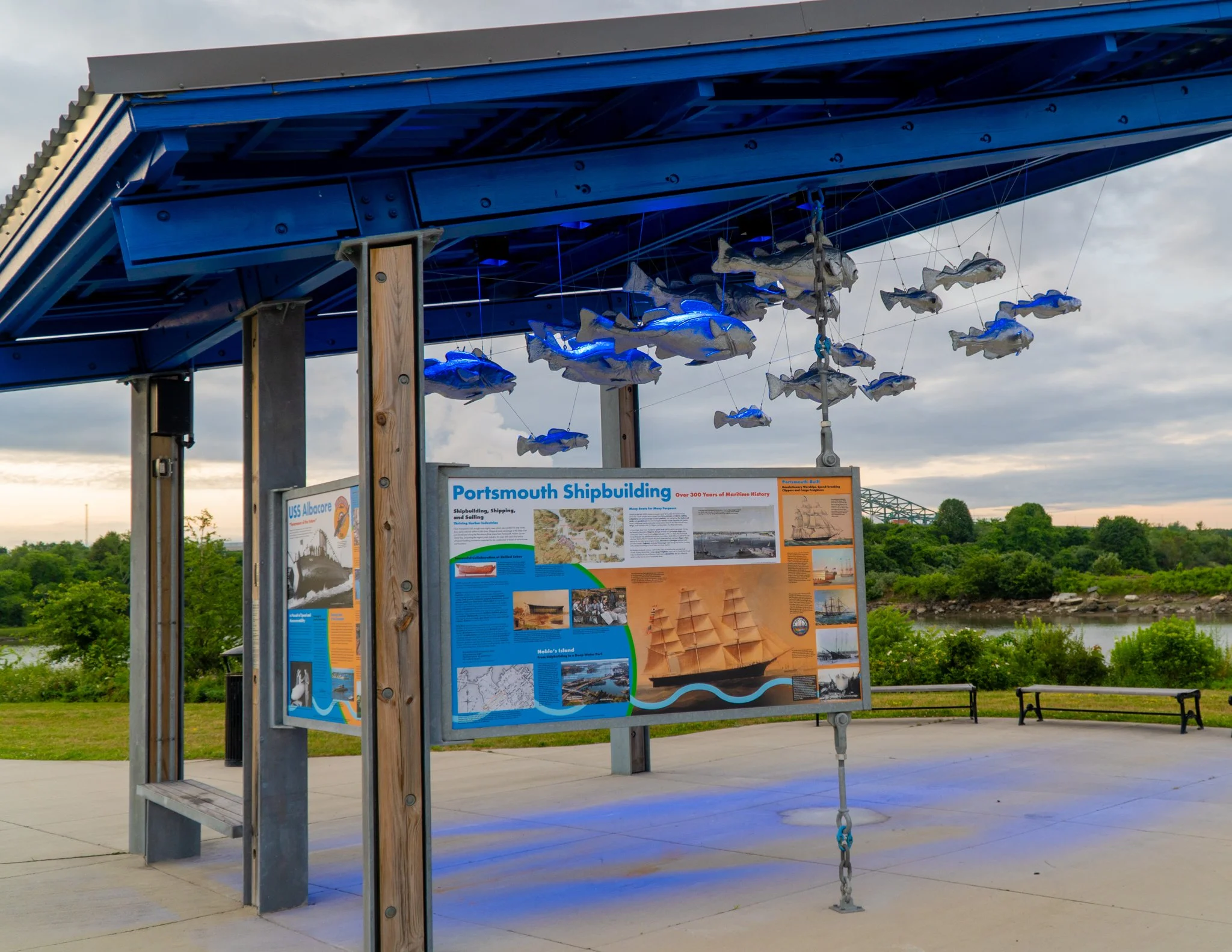 Outdoor exhibit at Portsmouth Shipbuilding with information panels and floating fish sculptures under a blue canopy, overlooking a river and green landscape.