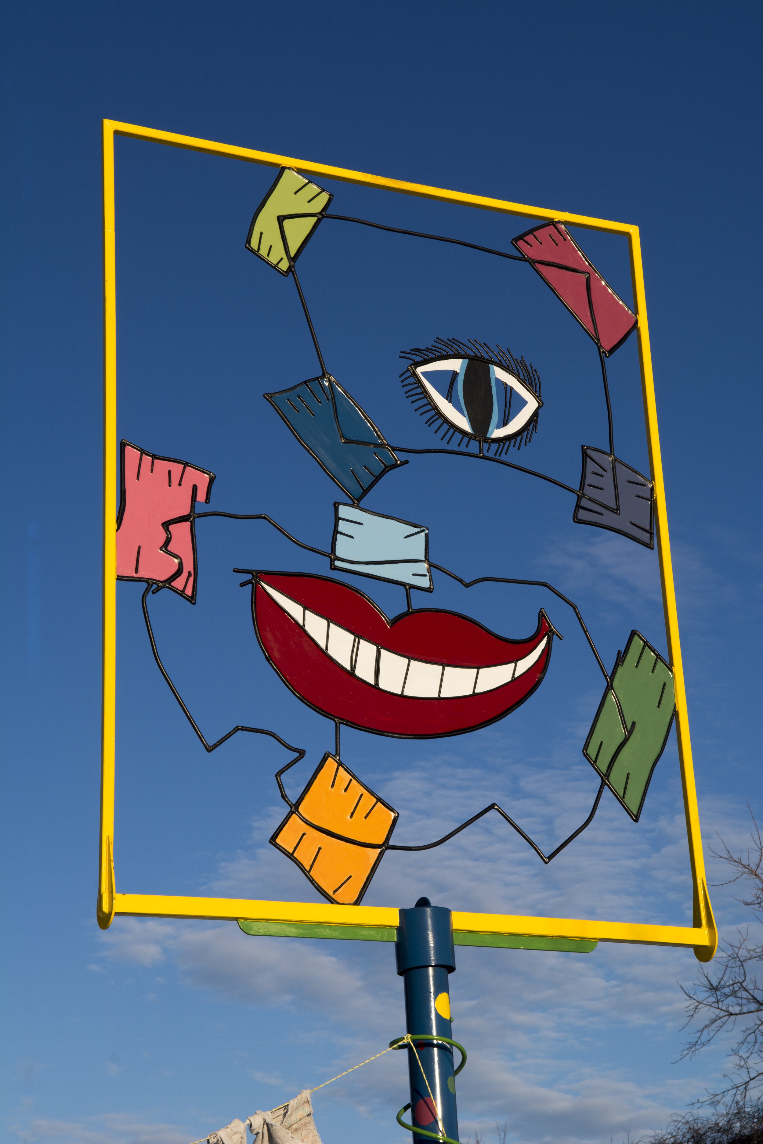 Colorful outdoor art installation featuring a smiling face with a blue eye, red lips, and multicolored geometric shapes on wires against a clear blue sky.