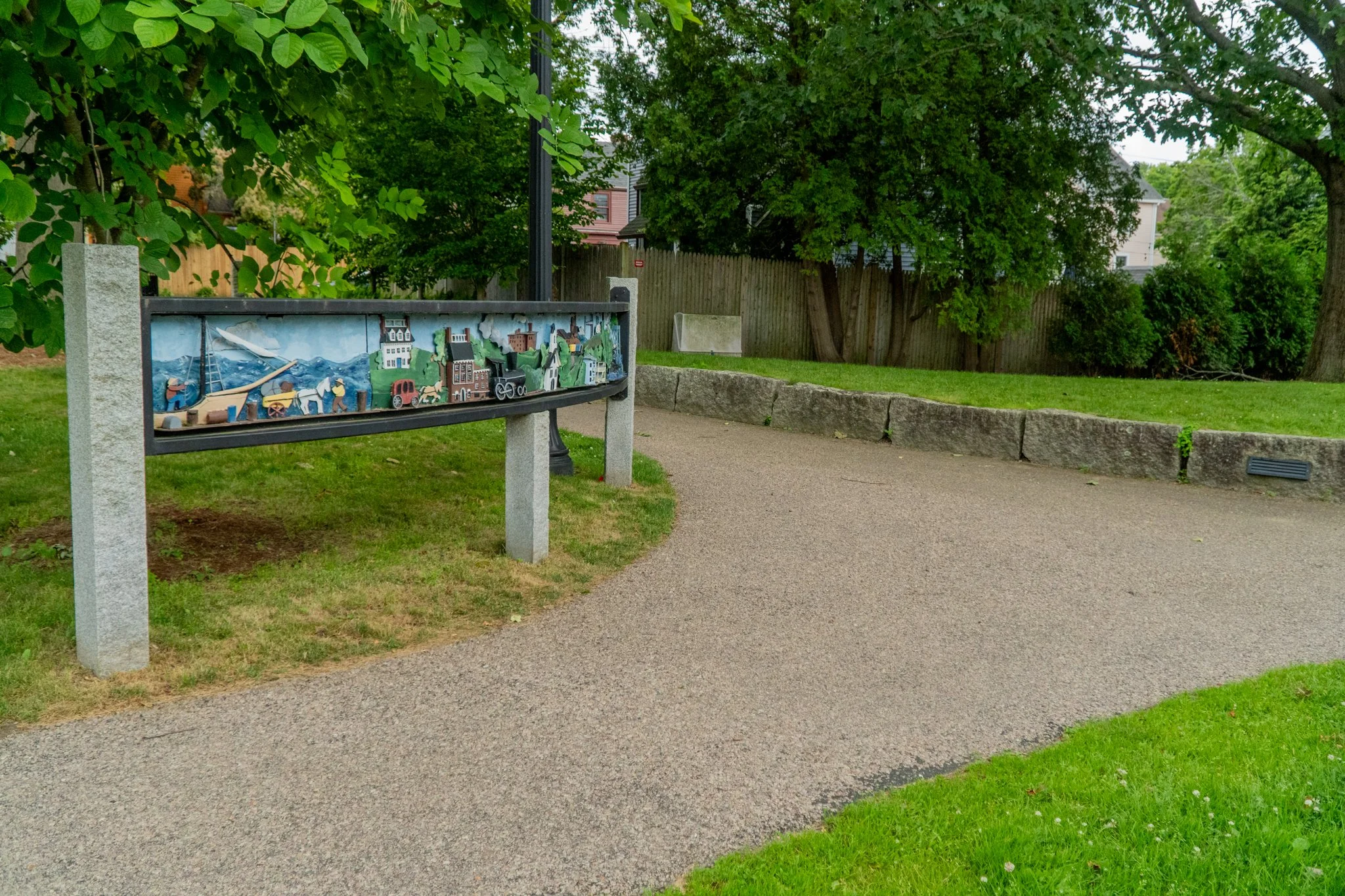 A park path with a colorful informational sign on the left, surrounded by green grass, trees, and a wooden fence in the background.