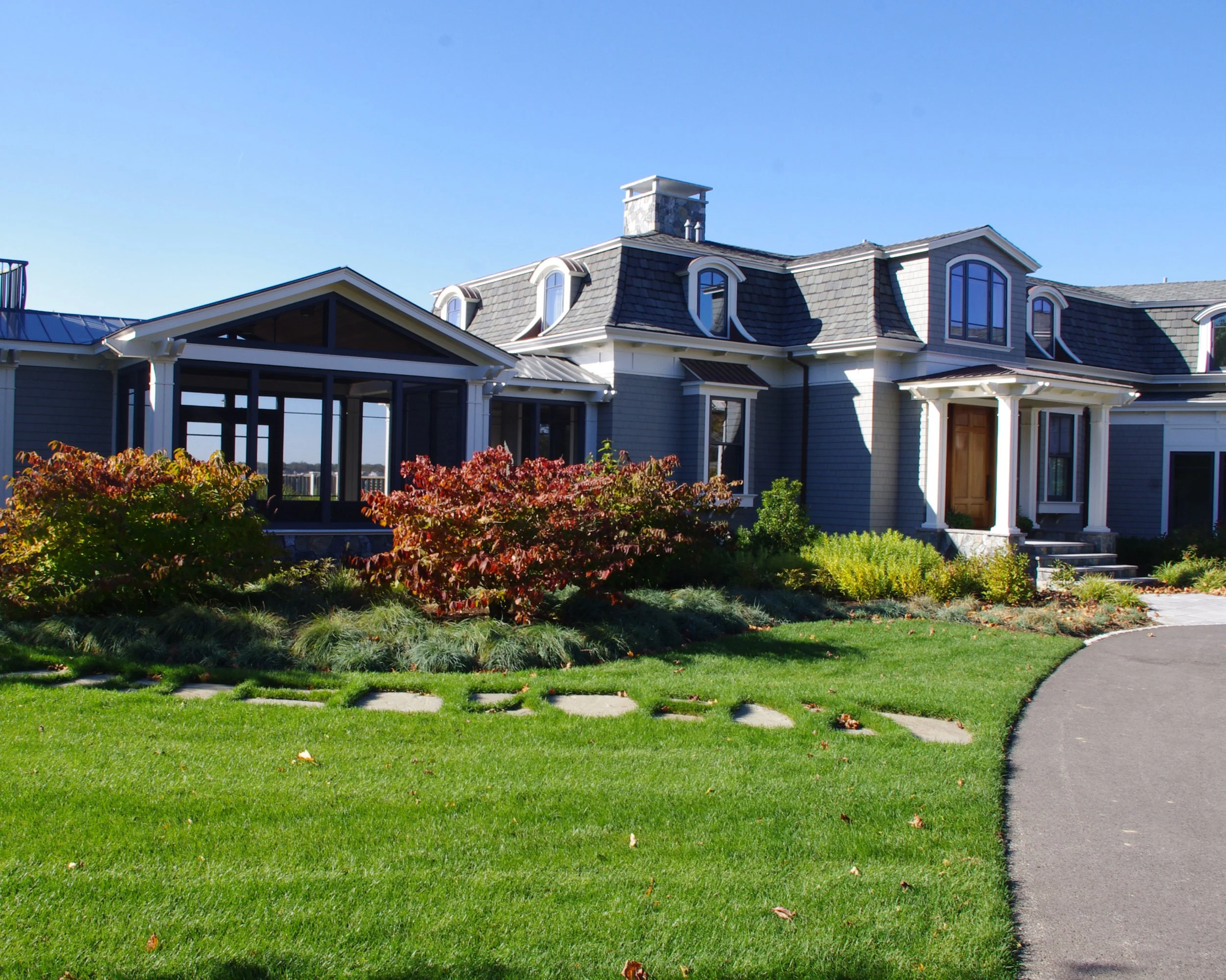 A large blue house with gray roof and multiple dormer windows, with white trim. There is a garden with shrubs and a lawn in front, along with a curved driveway.