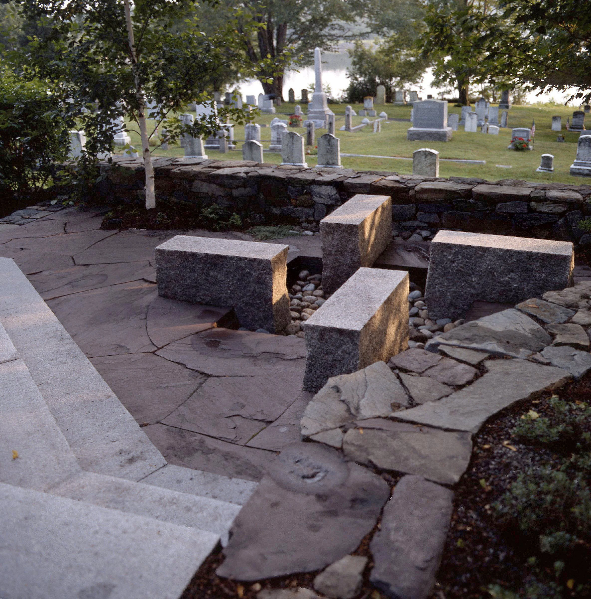 A stone pathway with steps leading to a memorial stone circle, with a fire pit and trees in the background.
