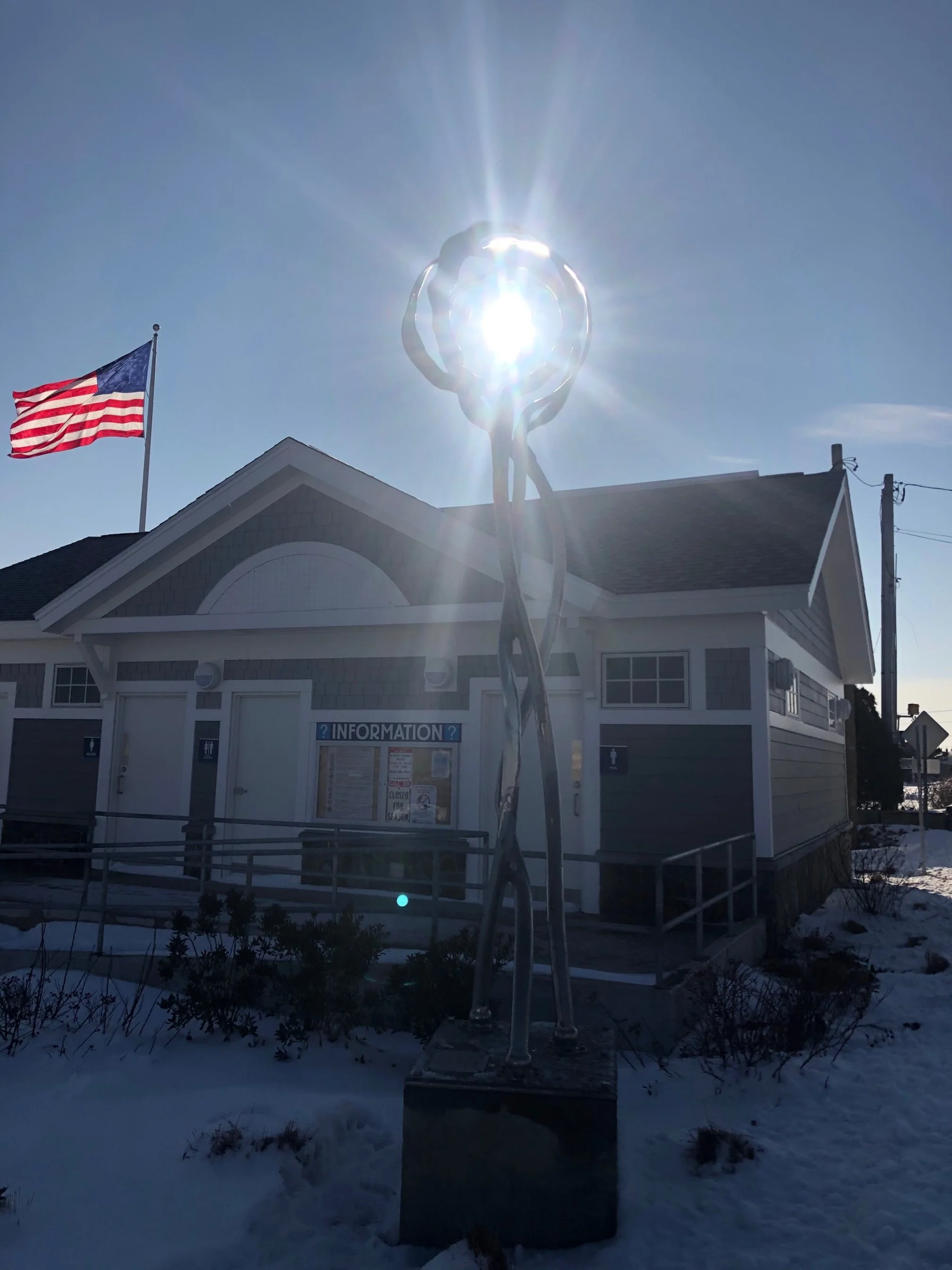 Sculpture with a sunburst effect behind it, located outside a building with an American flag and an information sign, on a snowy day.