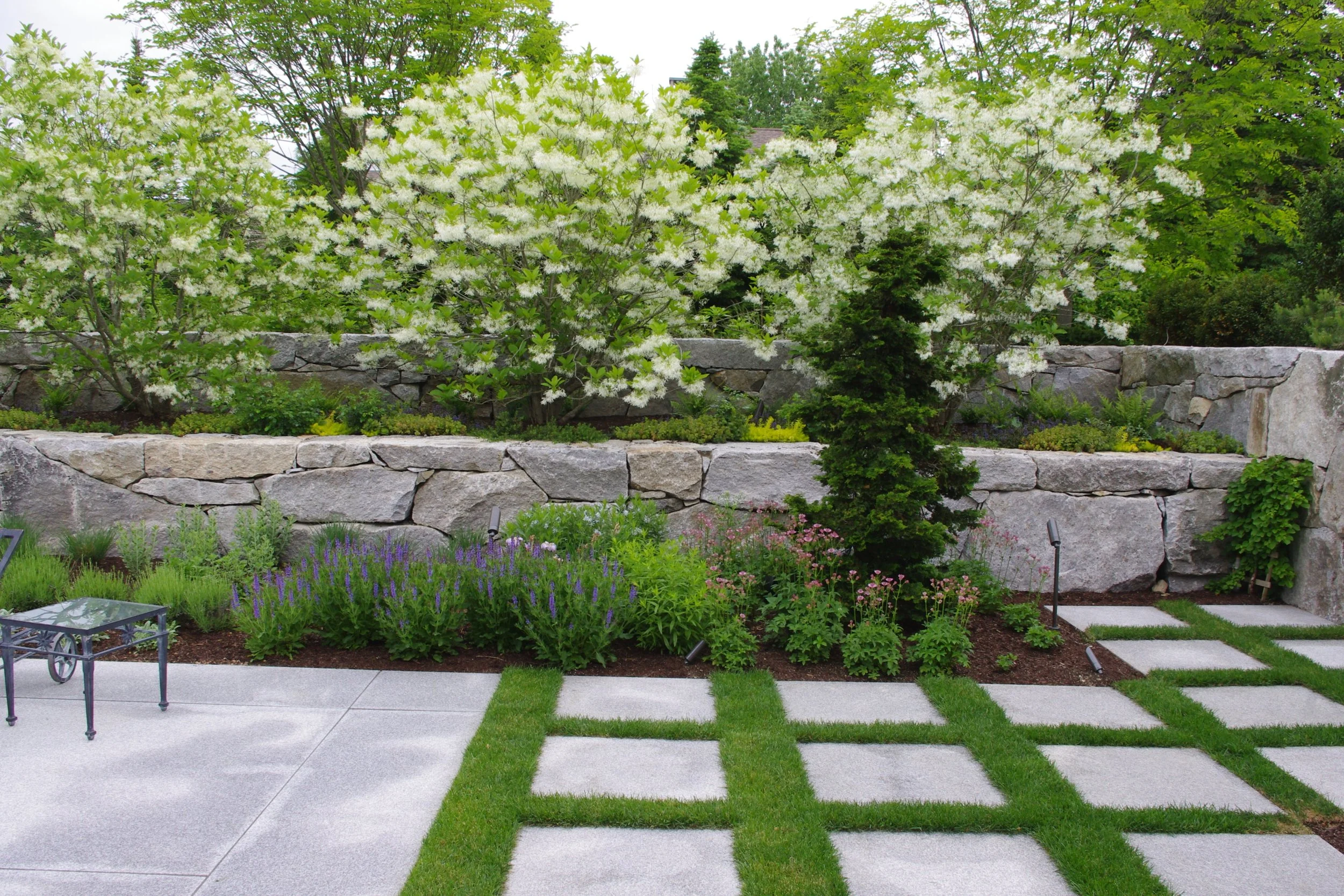 A landscaped backyard garden featuring a stone retaining wall, flowering trees, green shrubs, and a grassy area with a granite patio.
