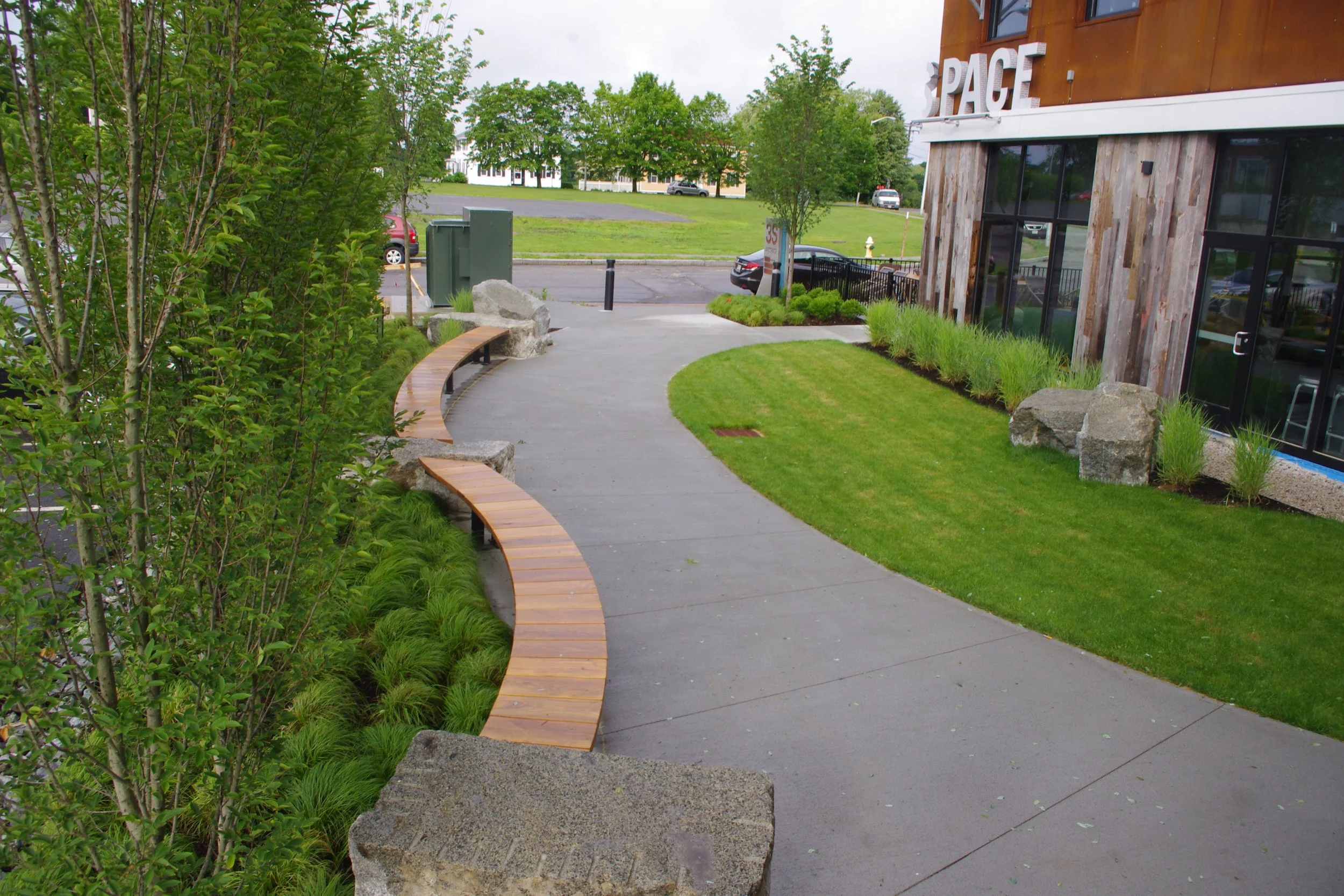 A sidewalk in front of a modern building with large windows, surrounded by green grass, shrubs, and rocks. There are curved wooden benches along the sidewalk, and a small area of landscaped plants and grass. In the background, a parking lot with cars