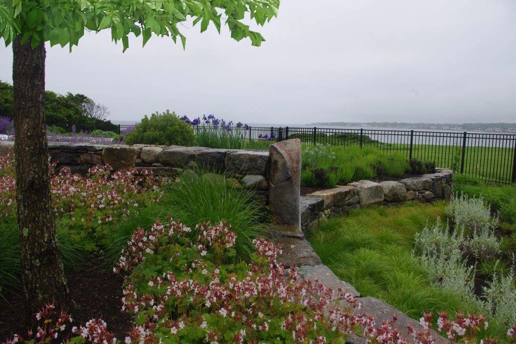A landscaped garden with pink and white flowers, green shrubs, and a stone wall by a black metal fence overlooking a body of water and a cloudy sky.