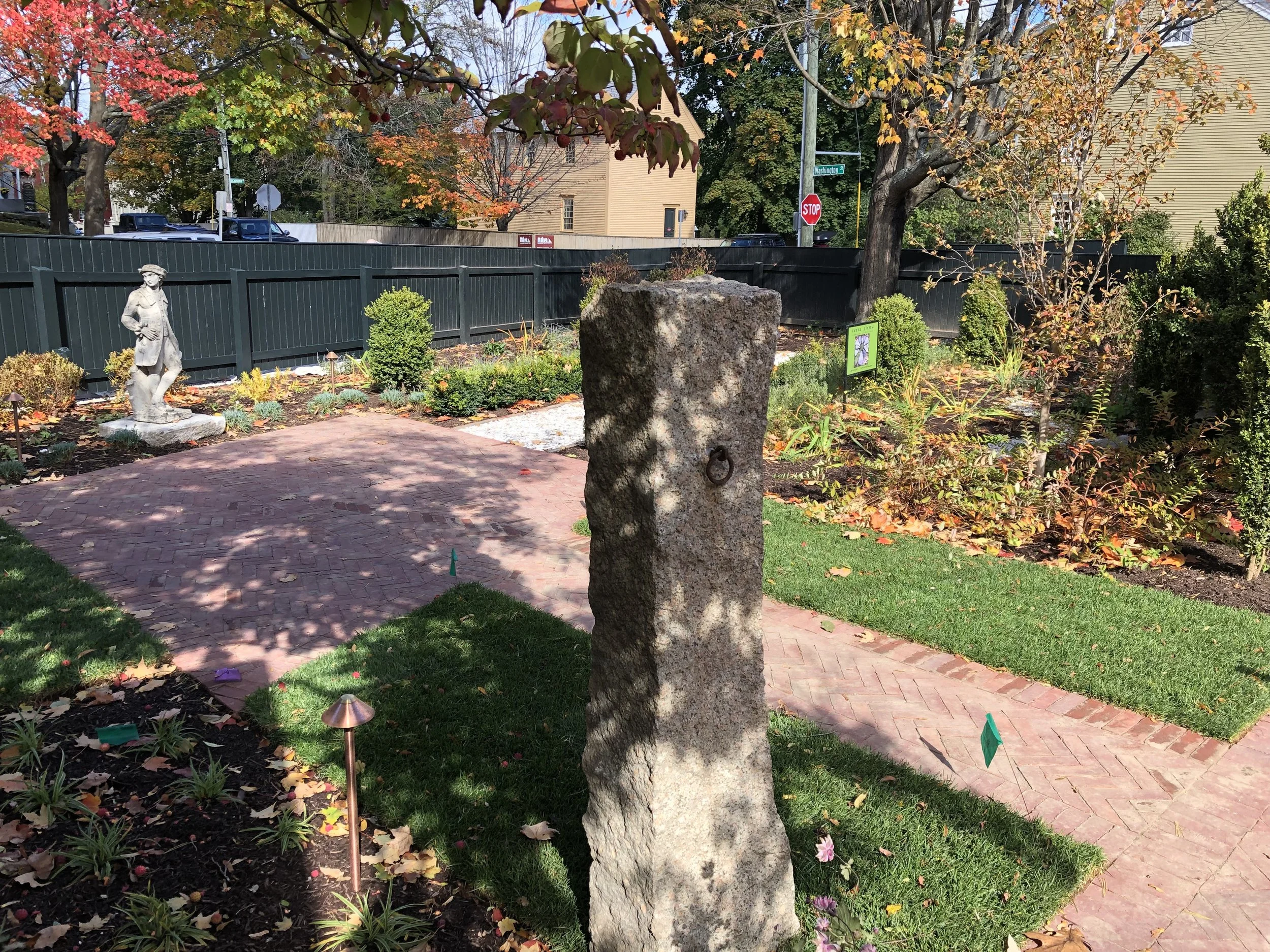 A small garden area with a stone sculpture of a person, a stone pillar with a ring, a brick pathway, and various plants and trees with autumn leaves.
