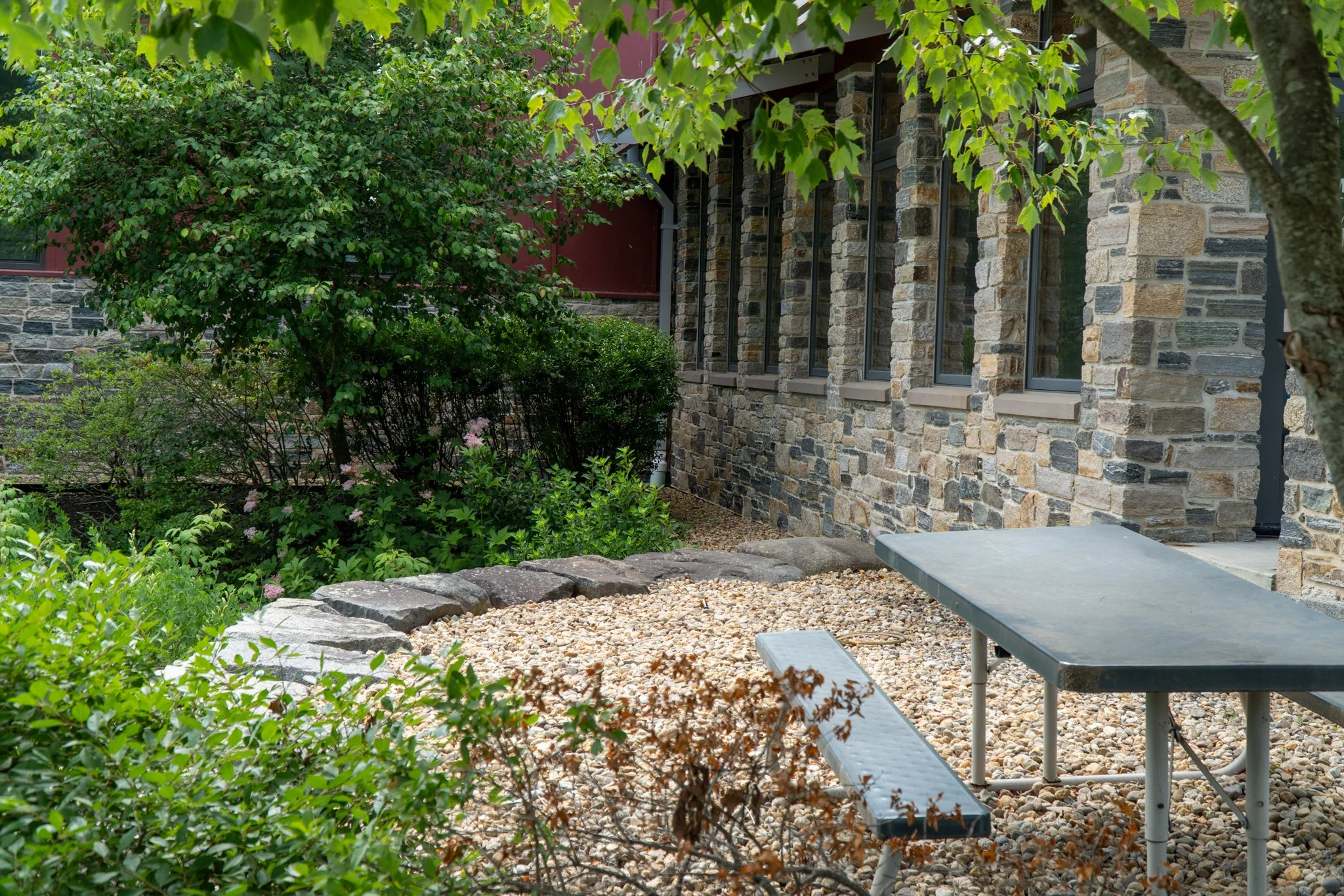 A stone building with large windows, surrounded by greenery including shrubs and trees, with a table and bench on a crushed stone surface in the foreground.