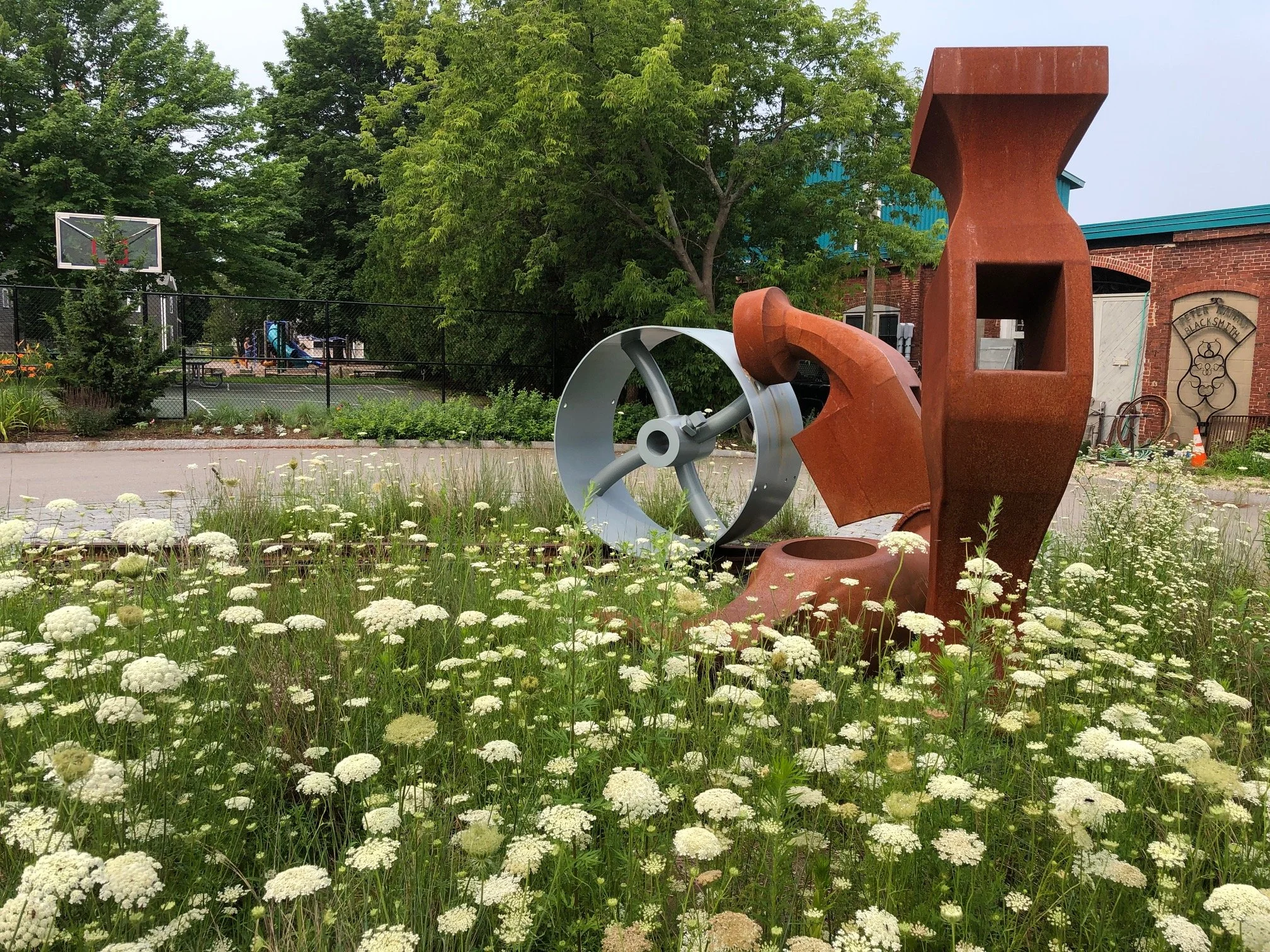 Outdoor art installation with metallic and rust-colored sculptures among wildflowers, located near a basketball court and a brick building.
