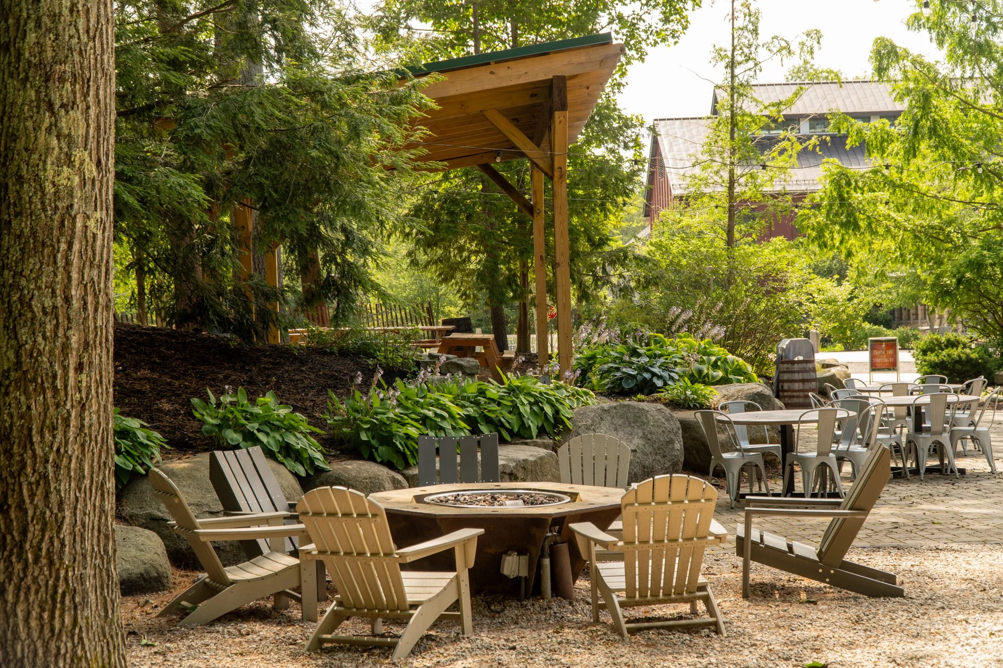 Outdoor seating area with Adirondack chairs and a fire pit surrounded by greenery and trees, with a picnic table under a wooden canopy.