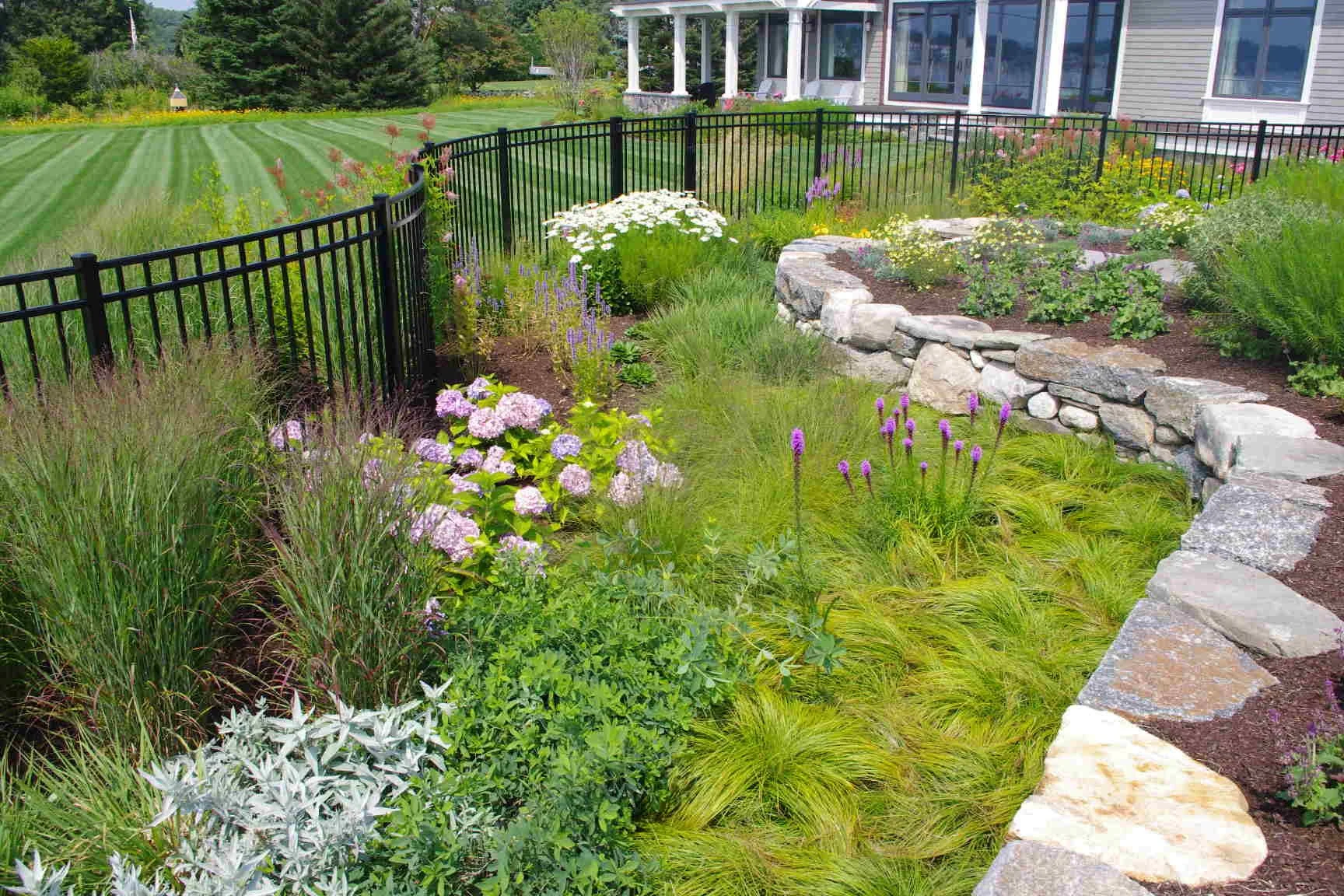 A landscaped garden with flowering plants, a stone wall, and a black metal fence, adjacent to a house with a patio, on a sunny day.