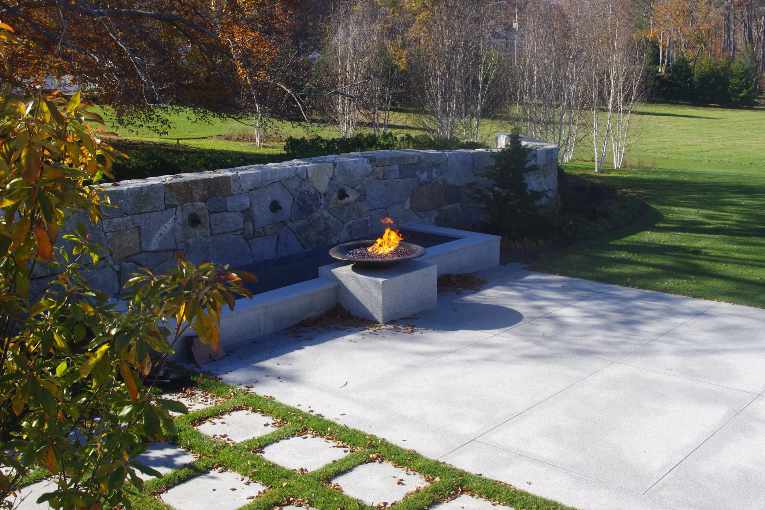Outdoor patio with a stone wall, a fire feature, granite pavers, and well-maintained grass, shrubs, and trees in the background.