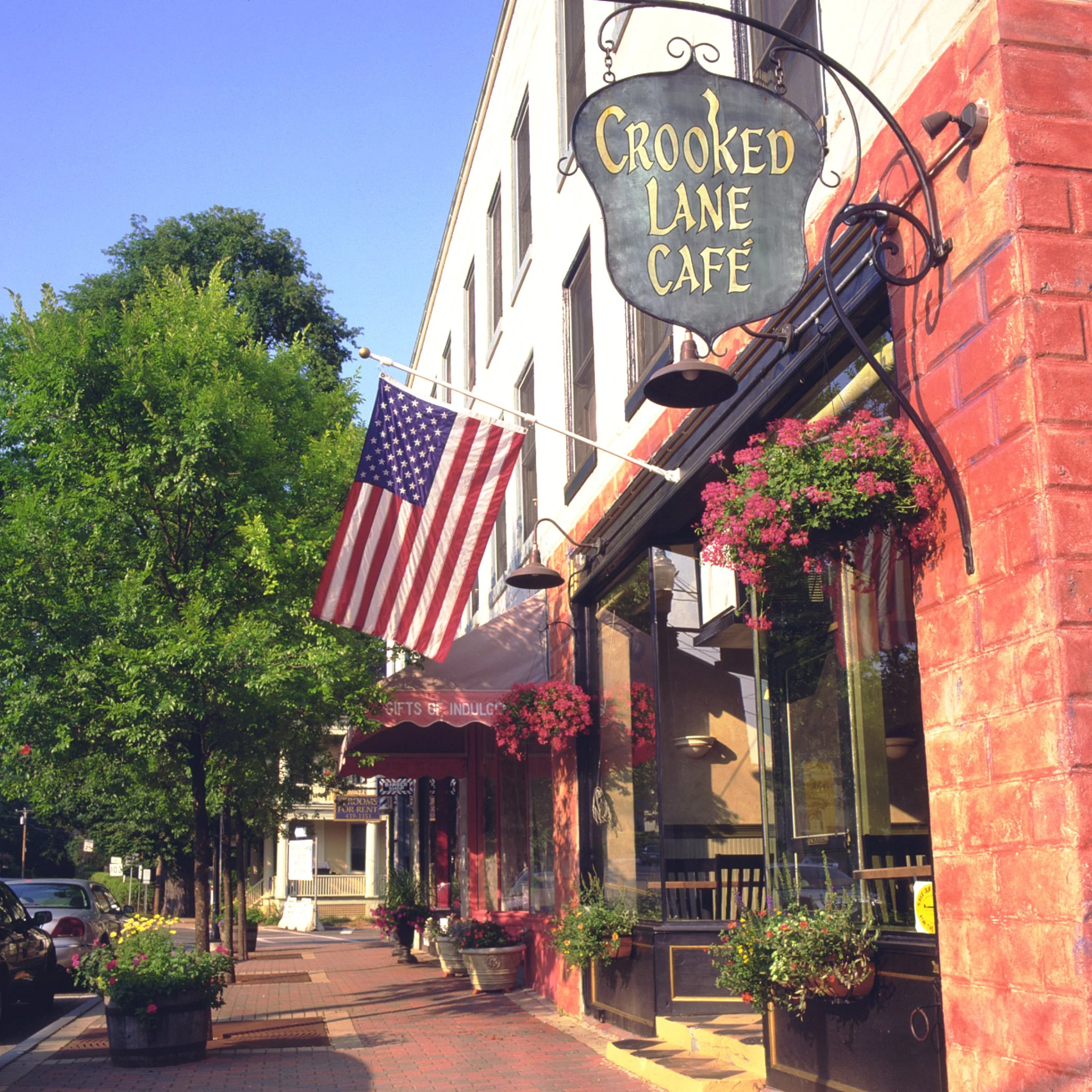 Exterior view of the Crooked Lane Café on a sunny day, featuring a hanging sign with the café's name, an American flag, potted flowers, and a brick sidewalk with a tree-lined street.