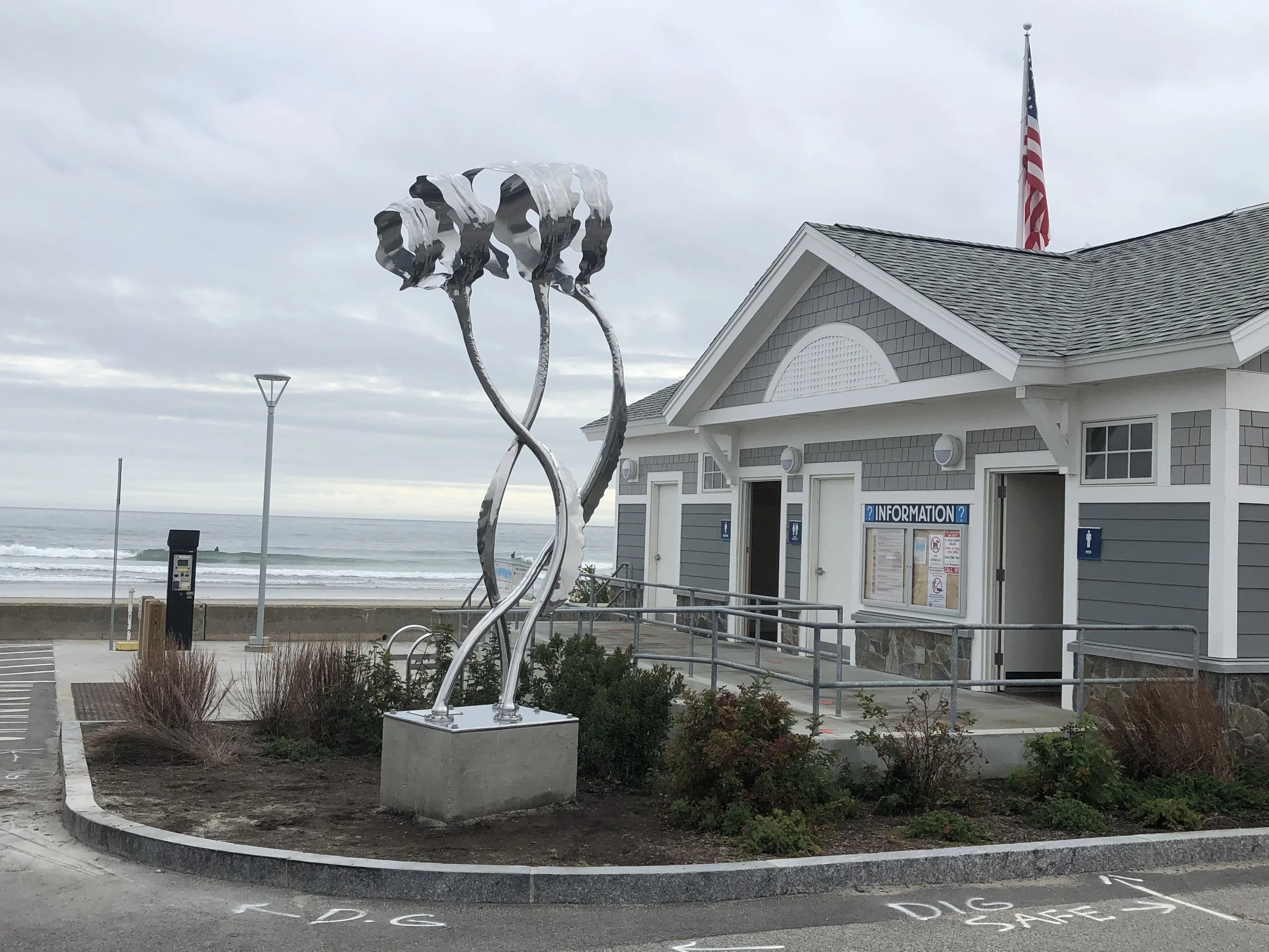 Public beach restroom building with a metal sculpture of intertwined kelp on a stone base in front, ocean in background, overcast sky, American flag flying.