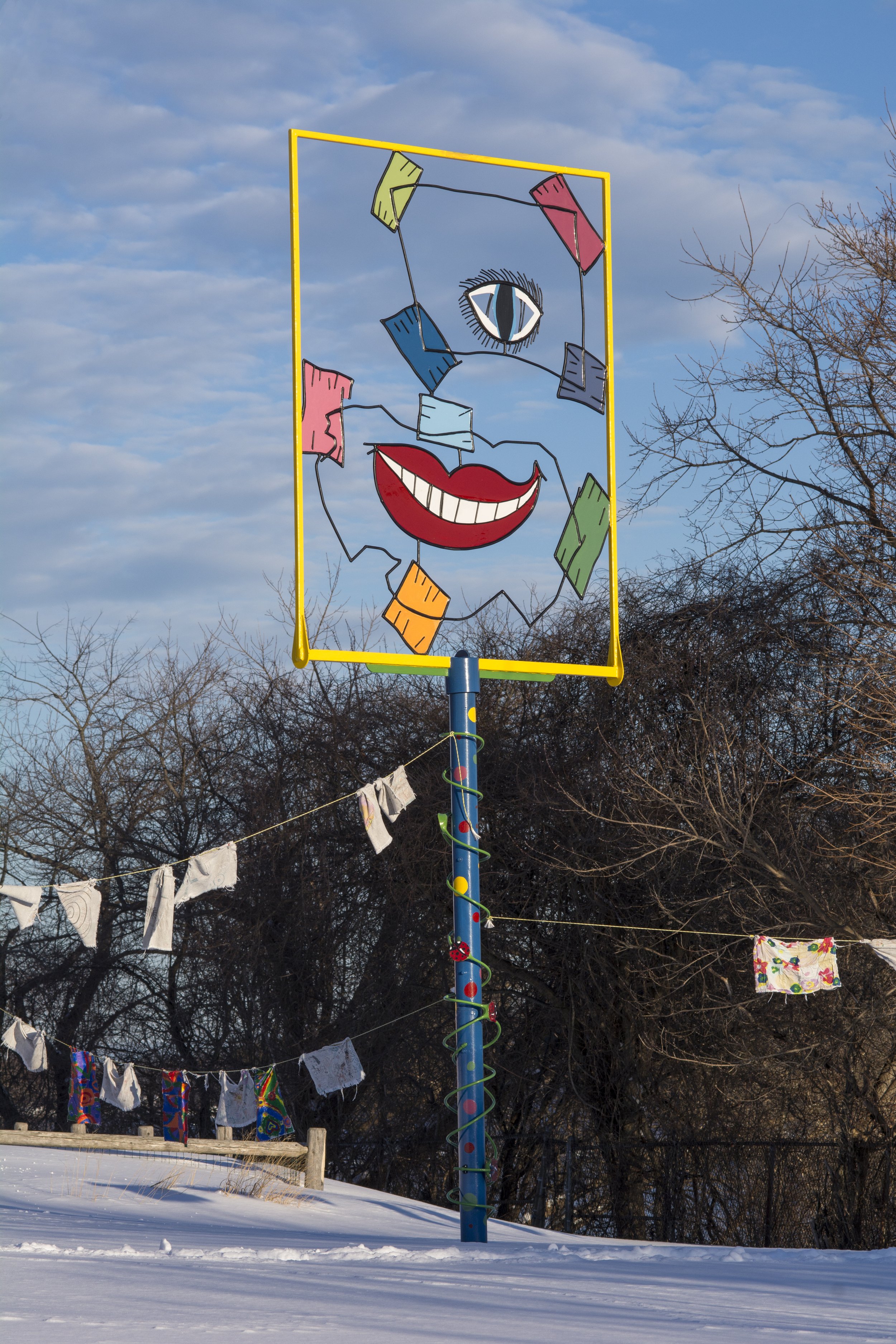 A colorful outdoor art installation of a smiling face with a single eye on a metal frame. The face has red lips, a wide white smile, and various brightly colored paper shapes around it. The sky in the background is partly cloudy and there are leafles