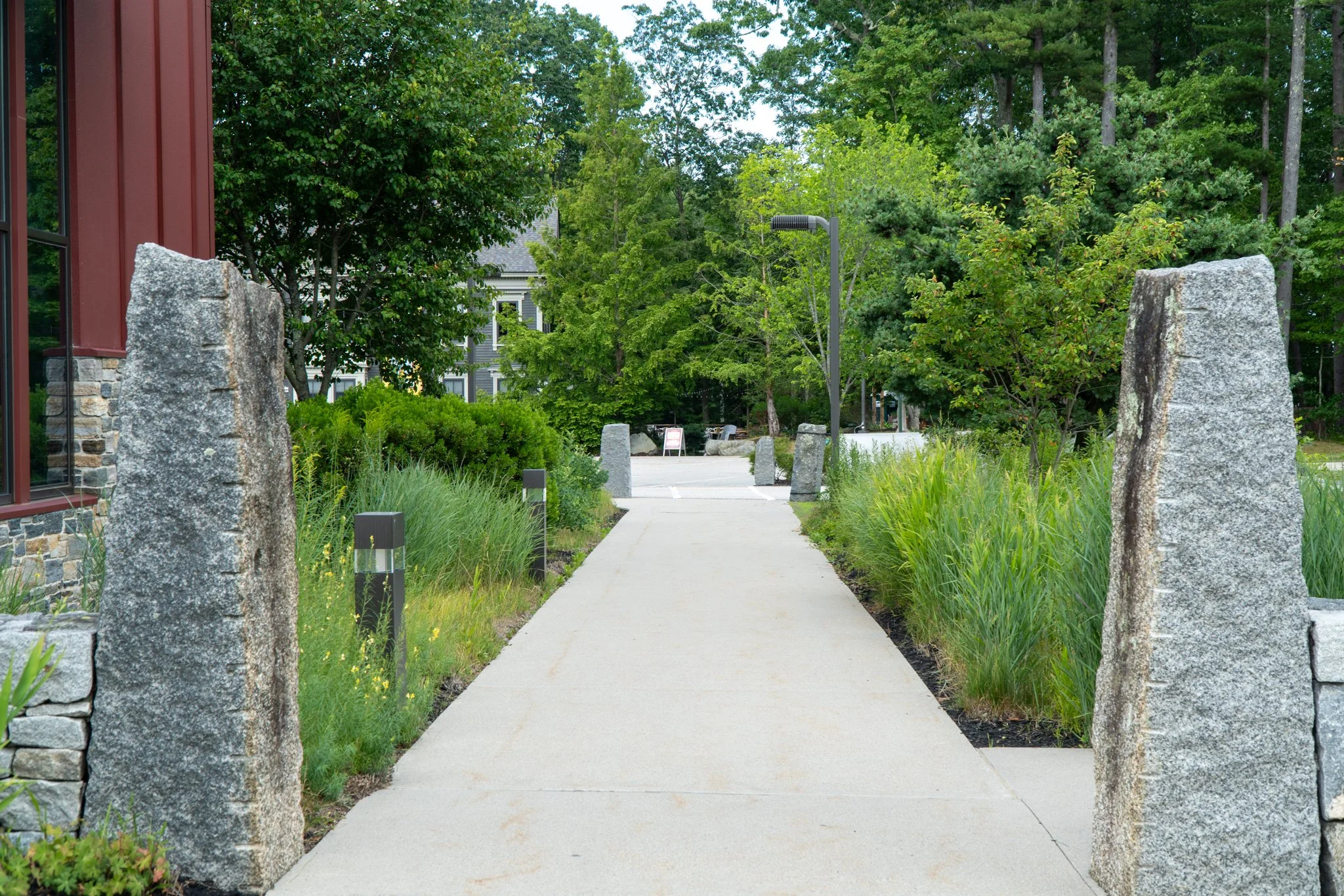 A concrete sidewalk flanked by large stone pillars and landscaped greenery leads to a gated entrance with a street and trees in the background.