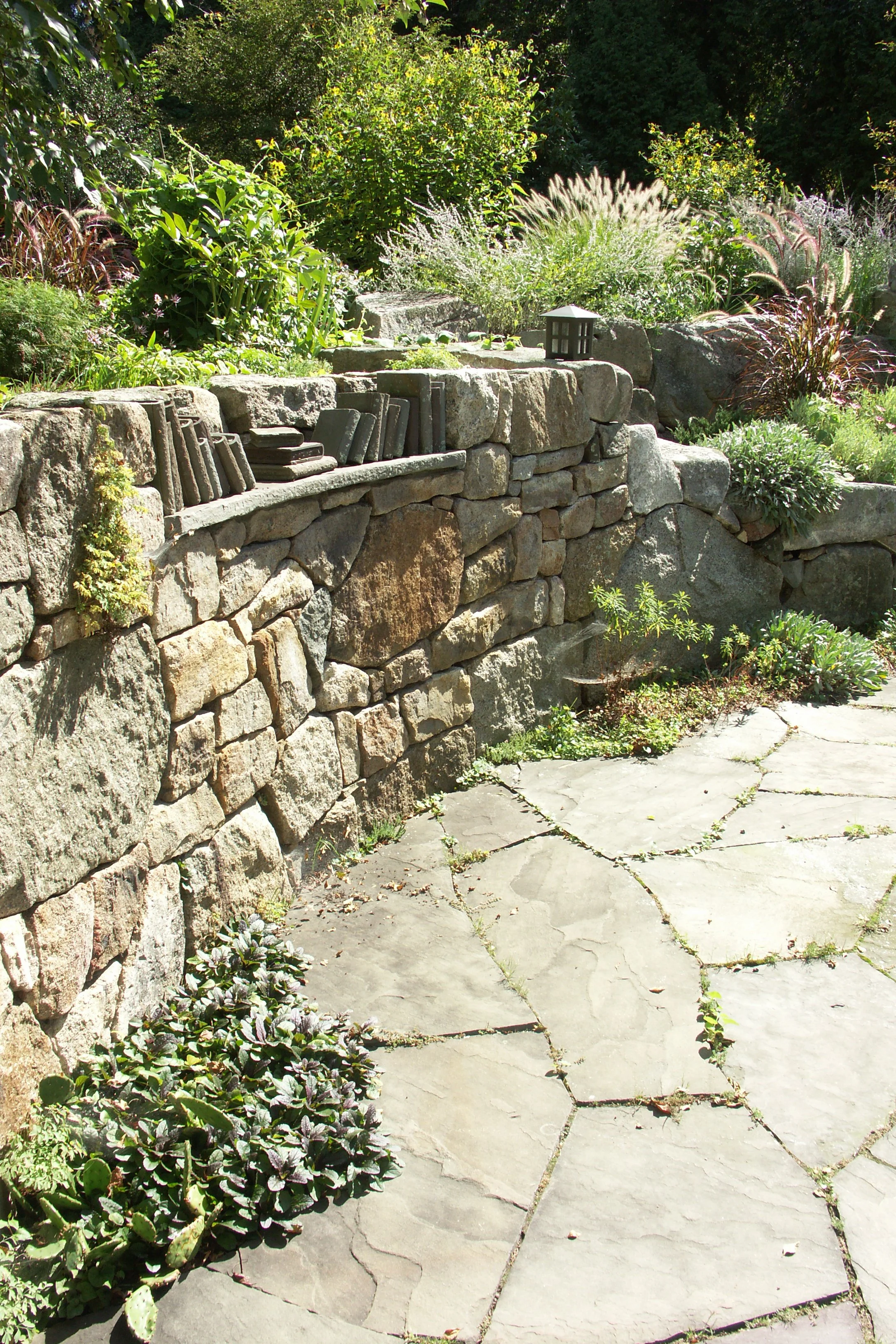 A stone patio with plants growing between cracks, a stone retaining wall with various plants and stone books, and a lush garden with tall grass and bushes in the background.