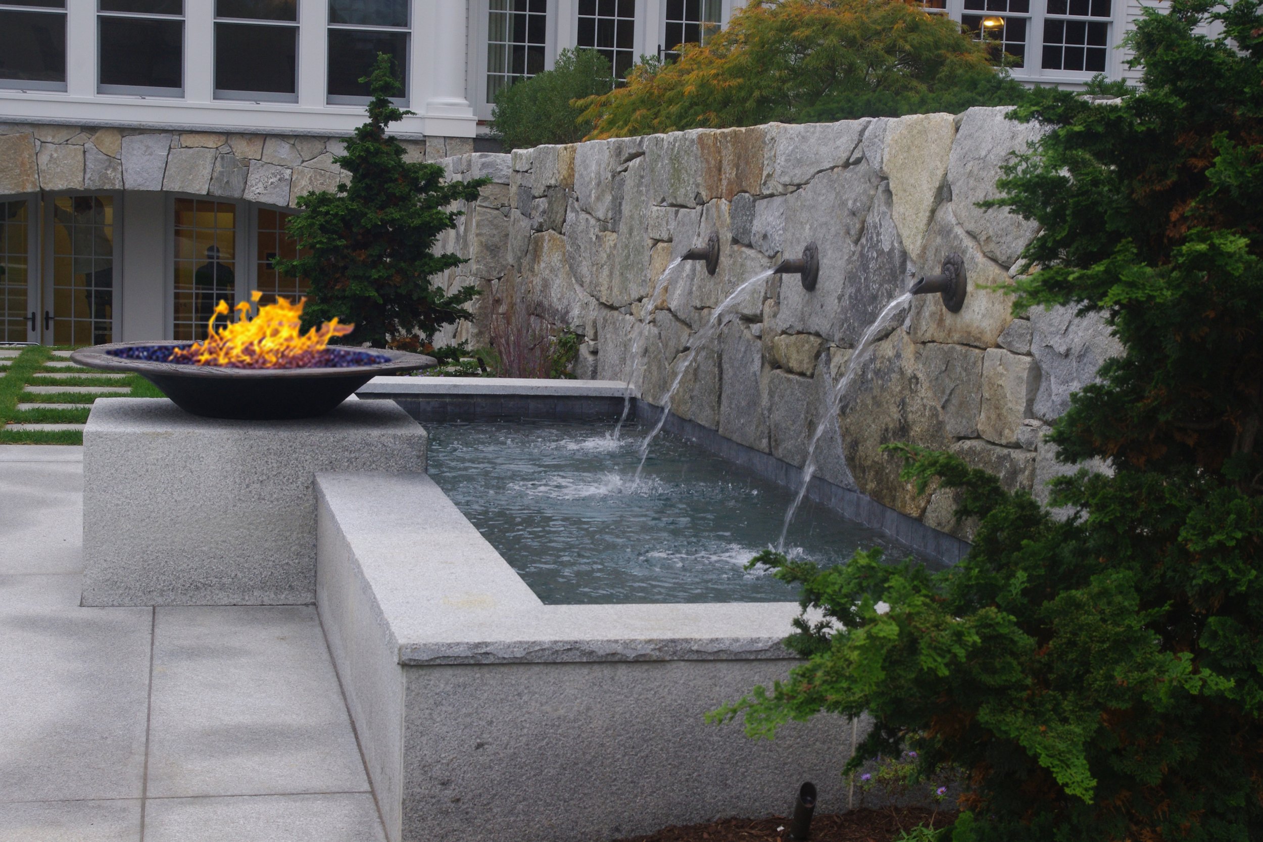 Outdoor water feature with a rectangular grey stone basin, a stone wall with three water spouts, and a fire bowl with flames in a landscaped garden area.