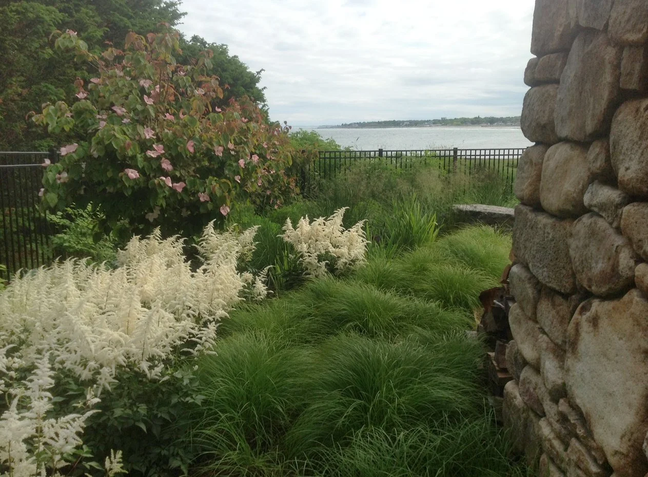 A garden view with lush green foliage, white and pink flowers, and a stone wall, overlooking a body of water with a distant shoreline under a cloudy sky.