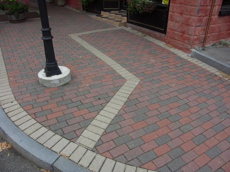 Brick sidewalk with a zigzag pattern outlined by lighter bricks near a street lamp post and building entrance.