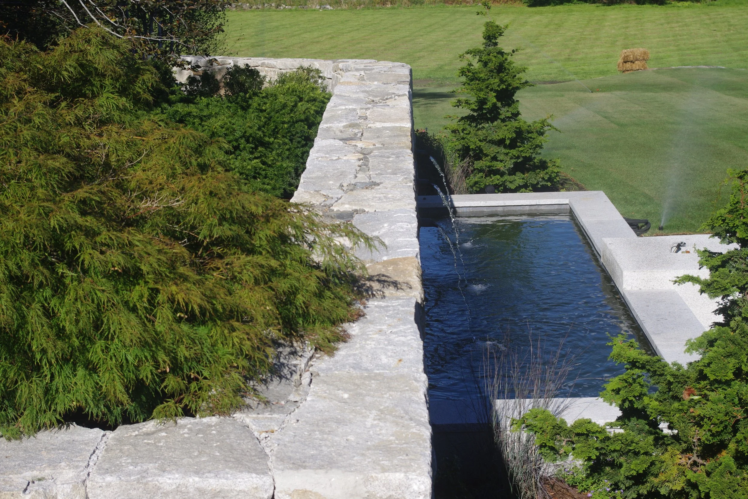 A stone wall with greenery on top and a water feature or small pool below, surrounded by trees and a grassy lawn.