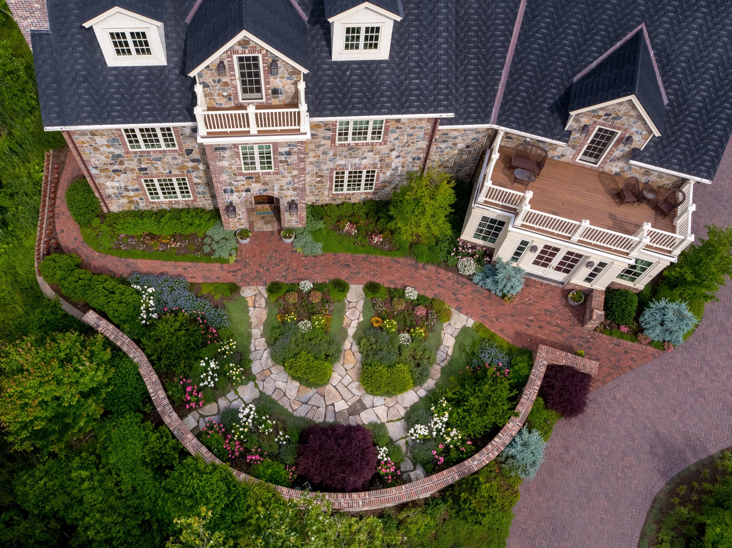 An aerial view of a large house with a stone facade, dark shingle roof, and multiple dormer windows. There are two levels of balconies on the house. A brick pathway and well-maintained garden with various plants, shrubs, and flowers surround the hous