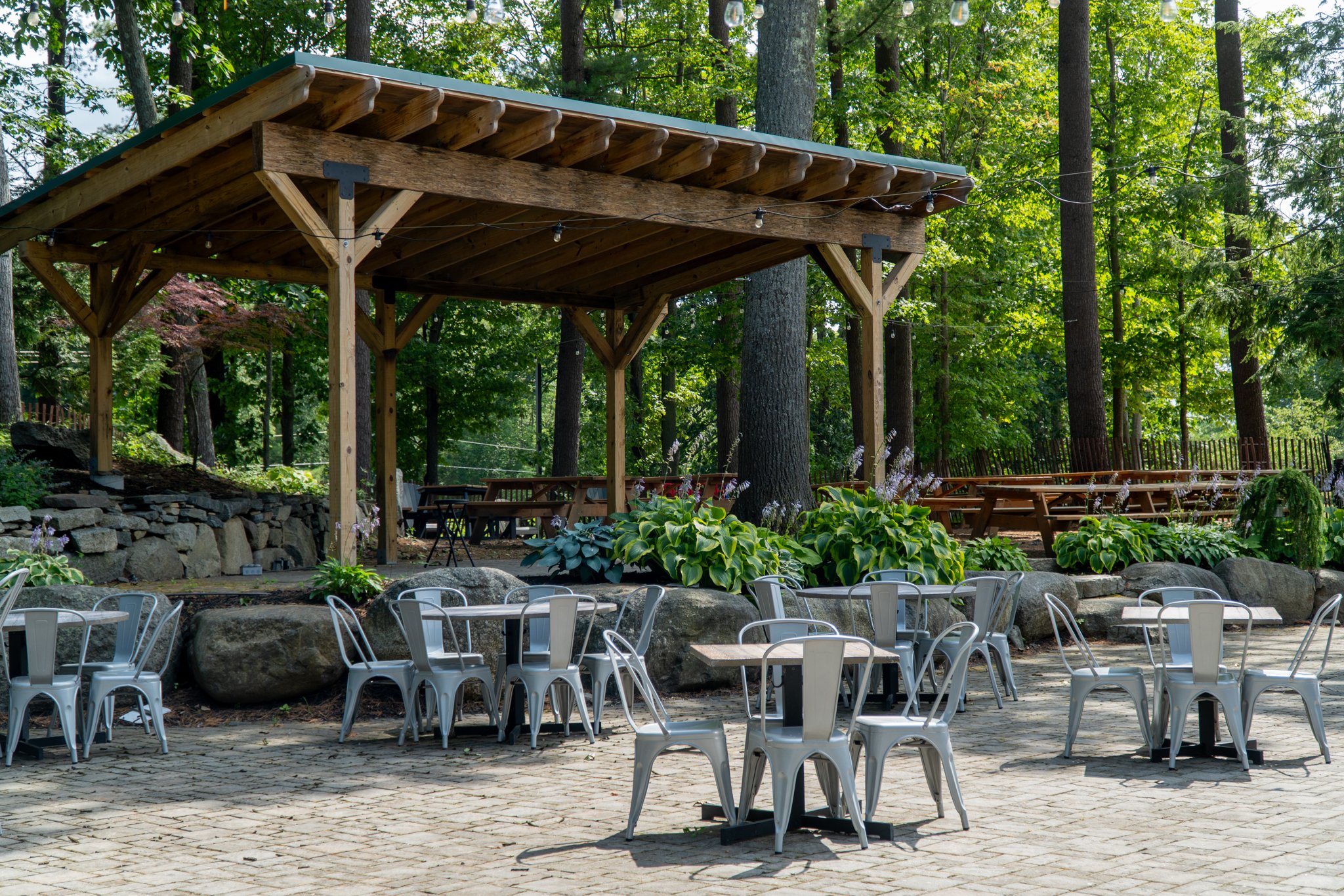 Outdoor patio with metal chairs and tables, a wooden pavilion, lush green trees and plants, and a stone wall in a forest setting.