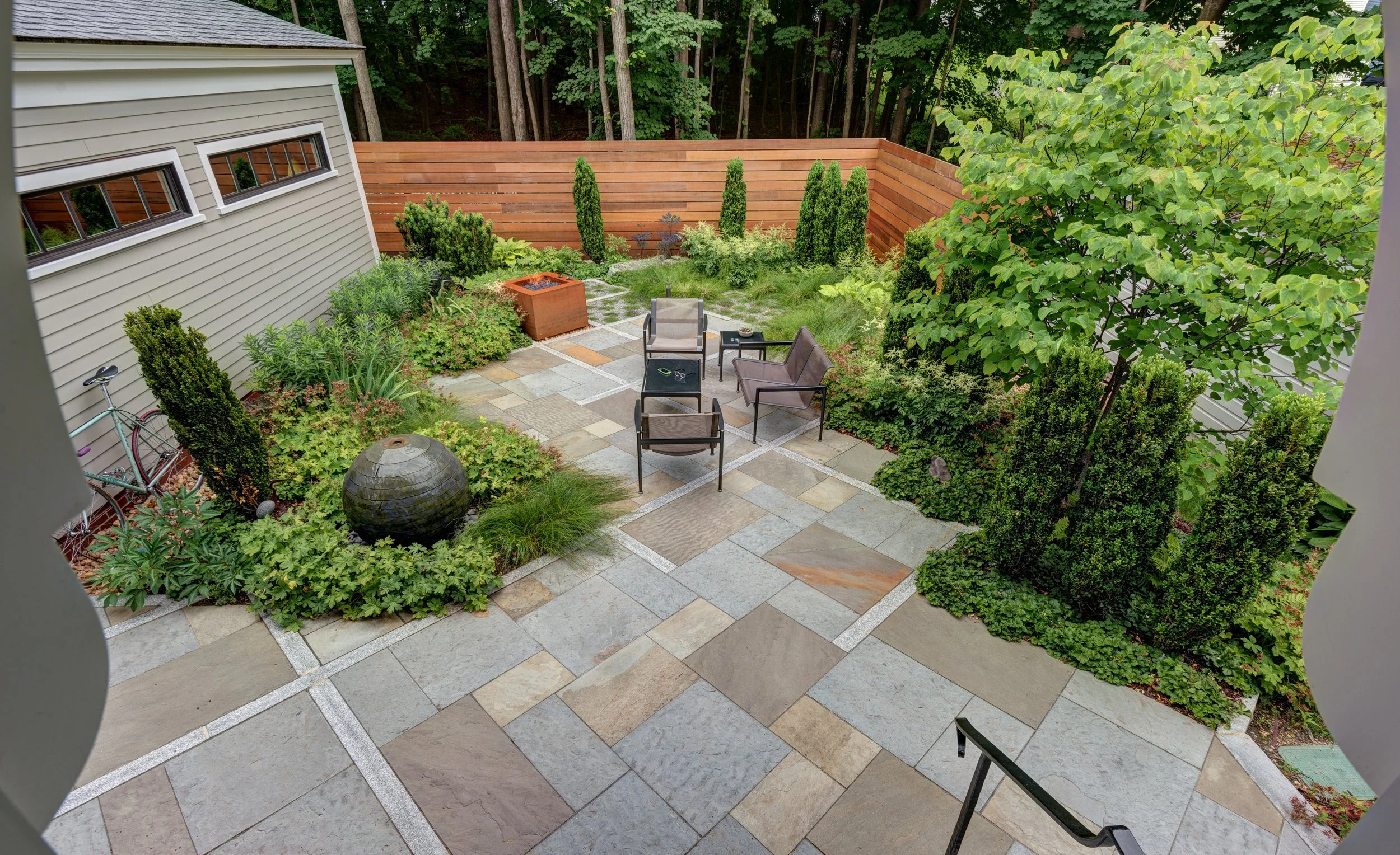 View of a backyard patio with stone paving, garden furniture including chairs and a table, a decorative water fountain, lush green plants, trees, and a wooden privacy fence.