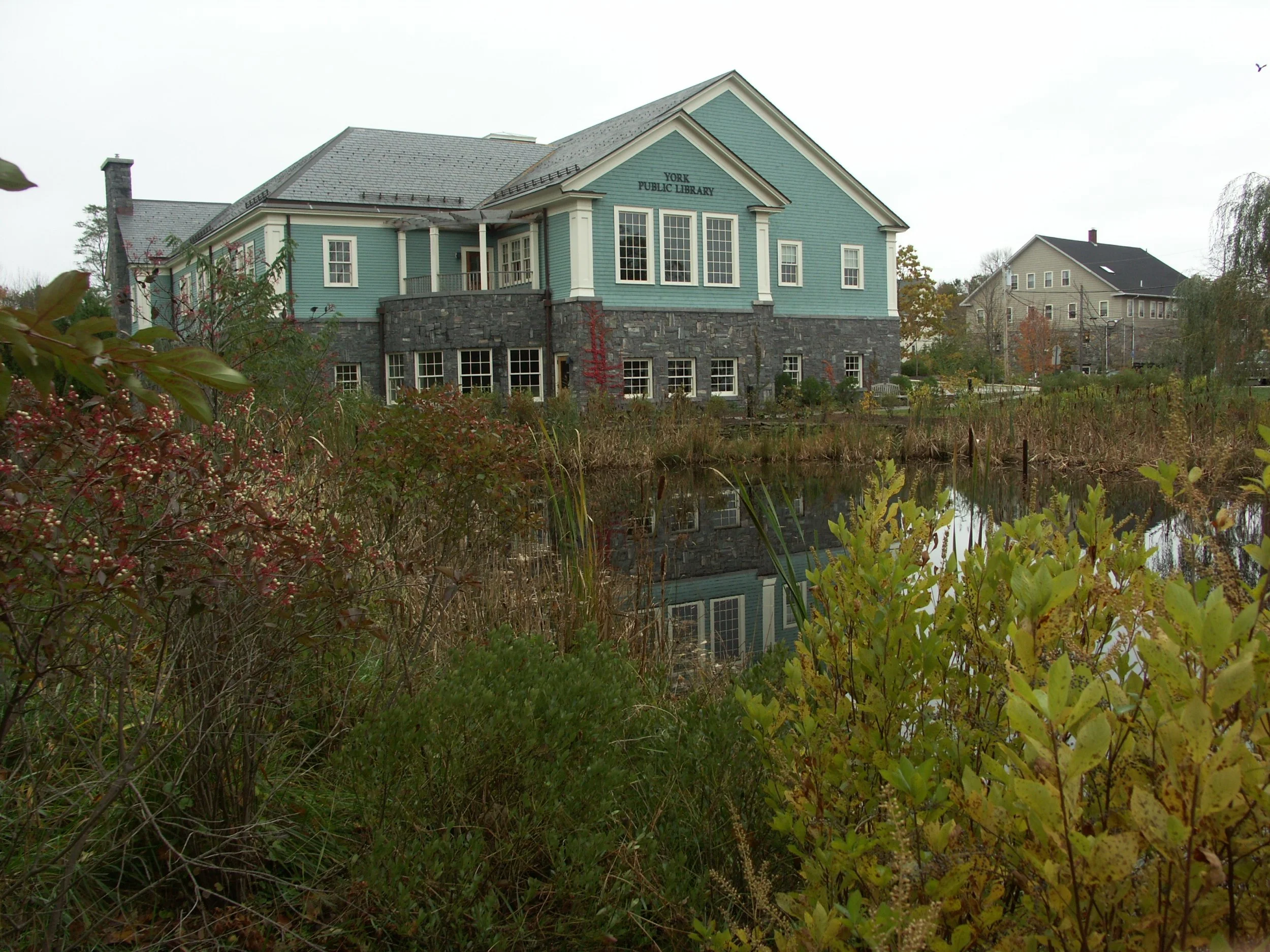 A large, blue, two-story building labeled 'York Public Library' with a gray stone foundation, surrounded by trees and shrubs near a pond.