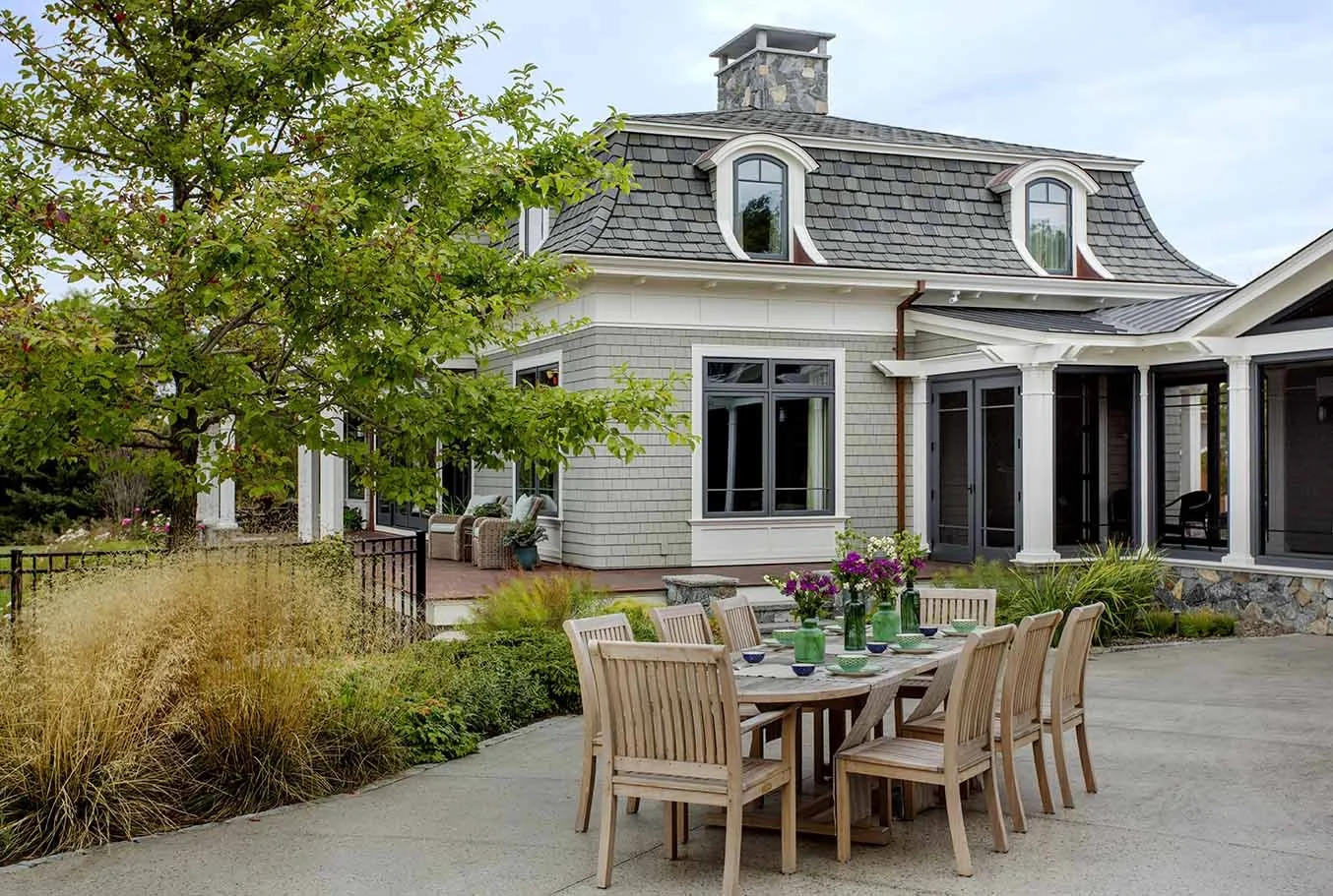 A backyard patio with a wooden dining table and chairs, surrounded by greenery and plants, in front of a two-story house with large windows and a shingled roof.