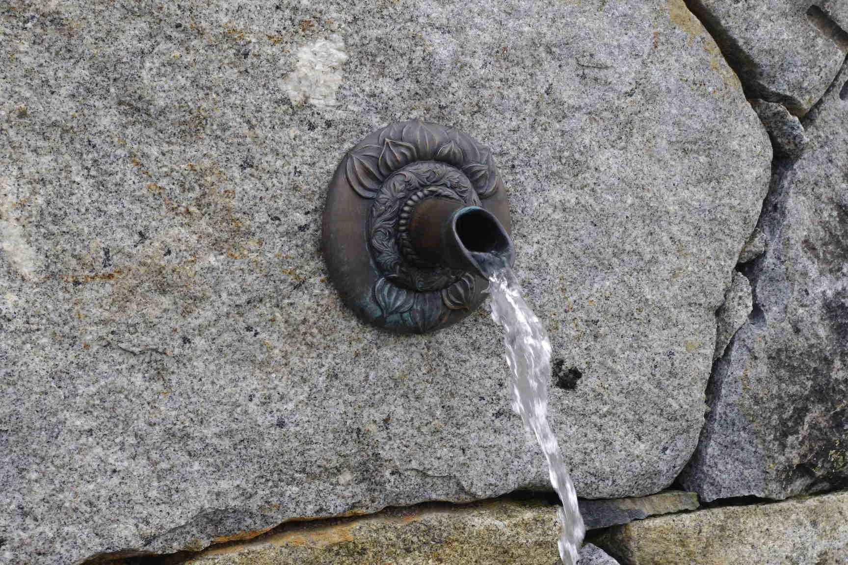 A decorative water fountain spout mounted on a stone wall, with water flowing out.