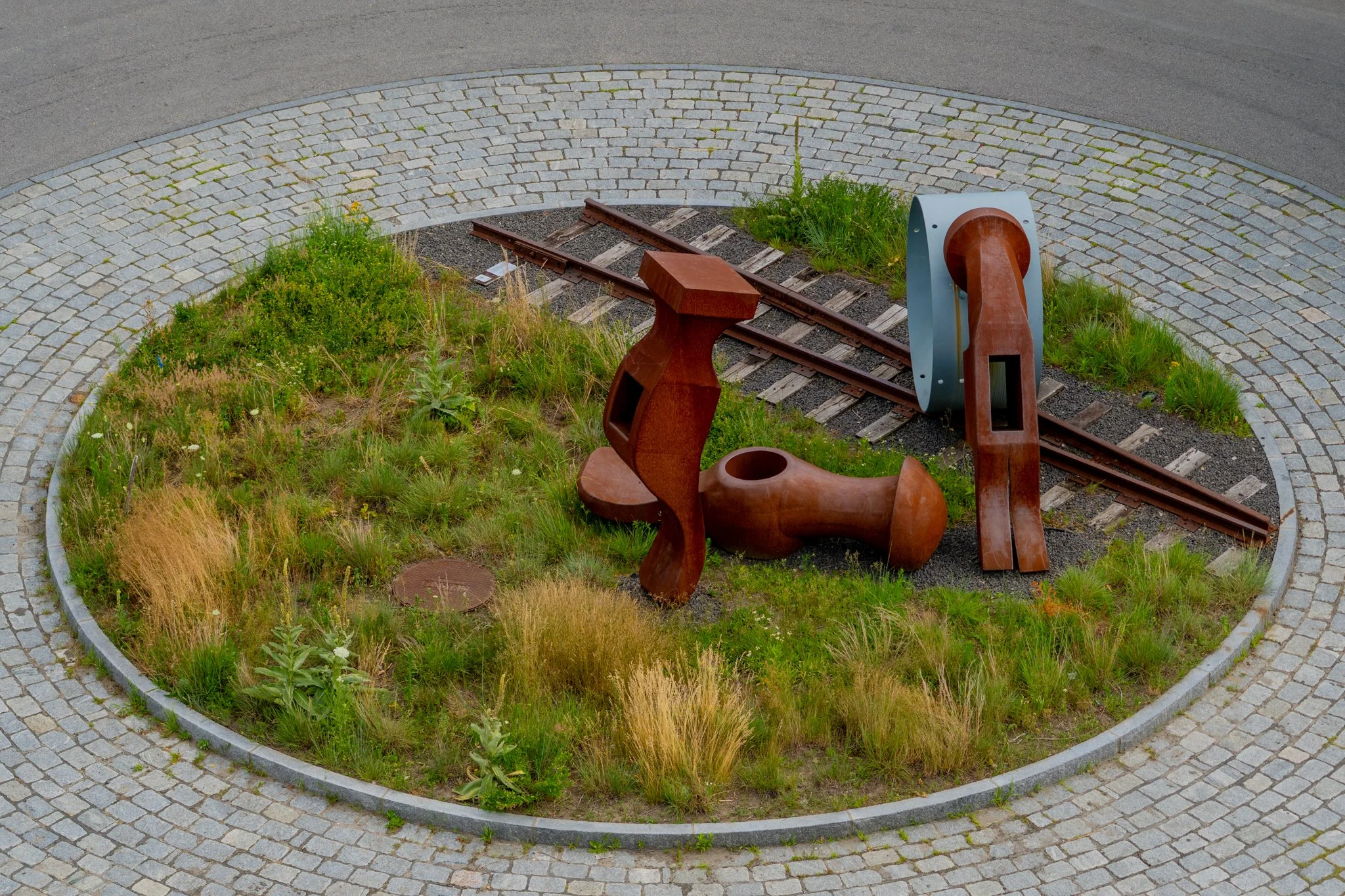 Outdoor art installation with industrial rust-colored sculptures, railway tracks, small plants, grass, and cobblestone circular pathway.