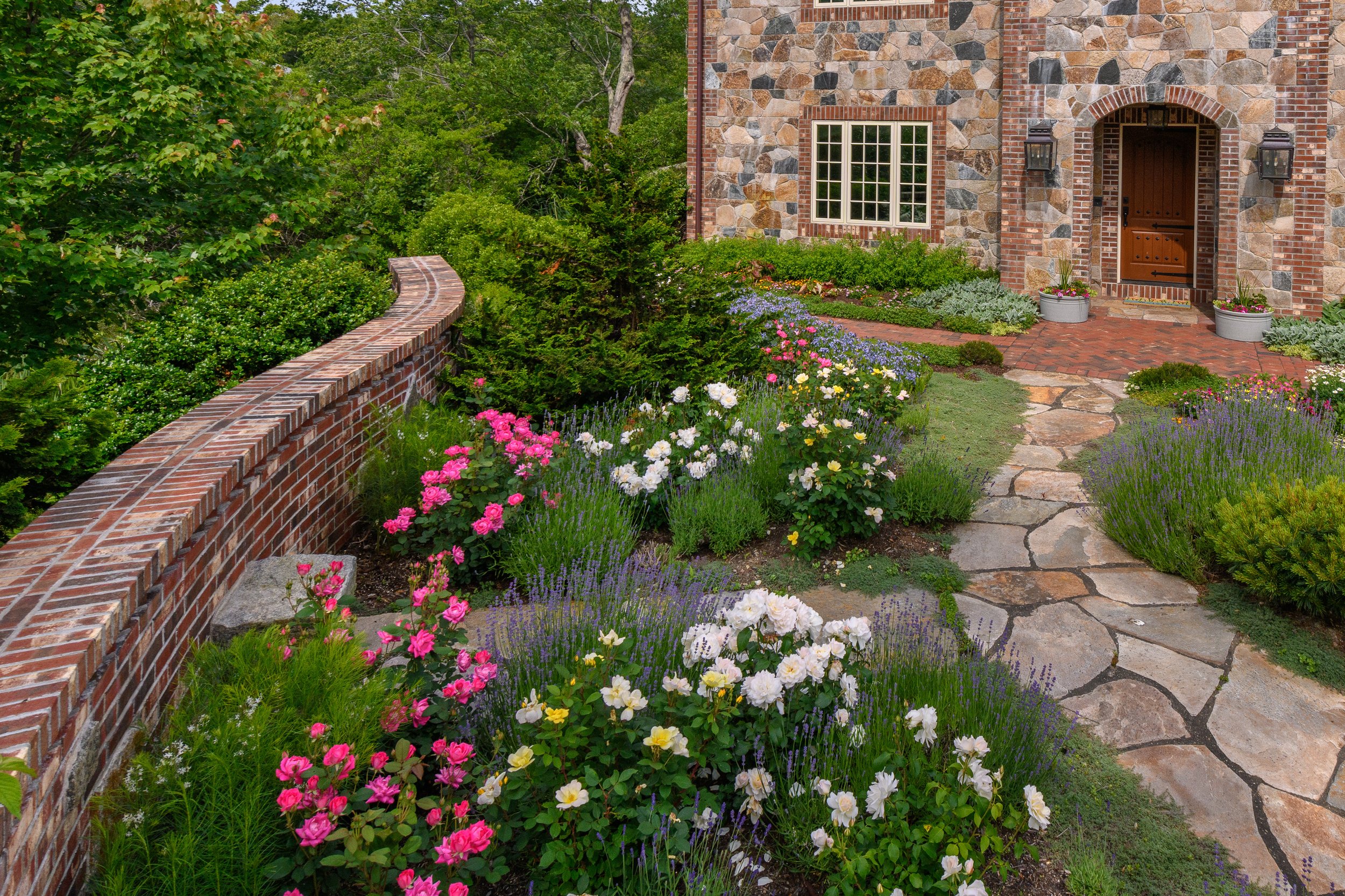Garden pathway leading to a brick house with a stone exterior, surrounded by colorful flowering plants and greenery.