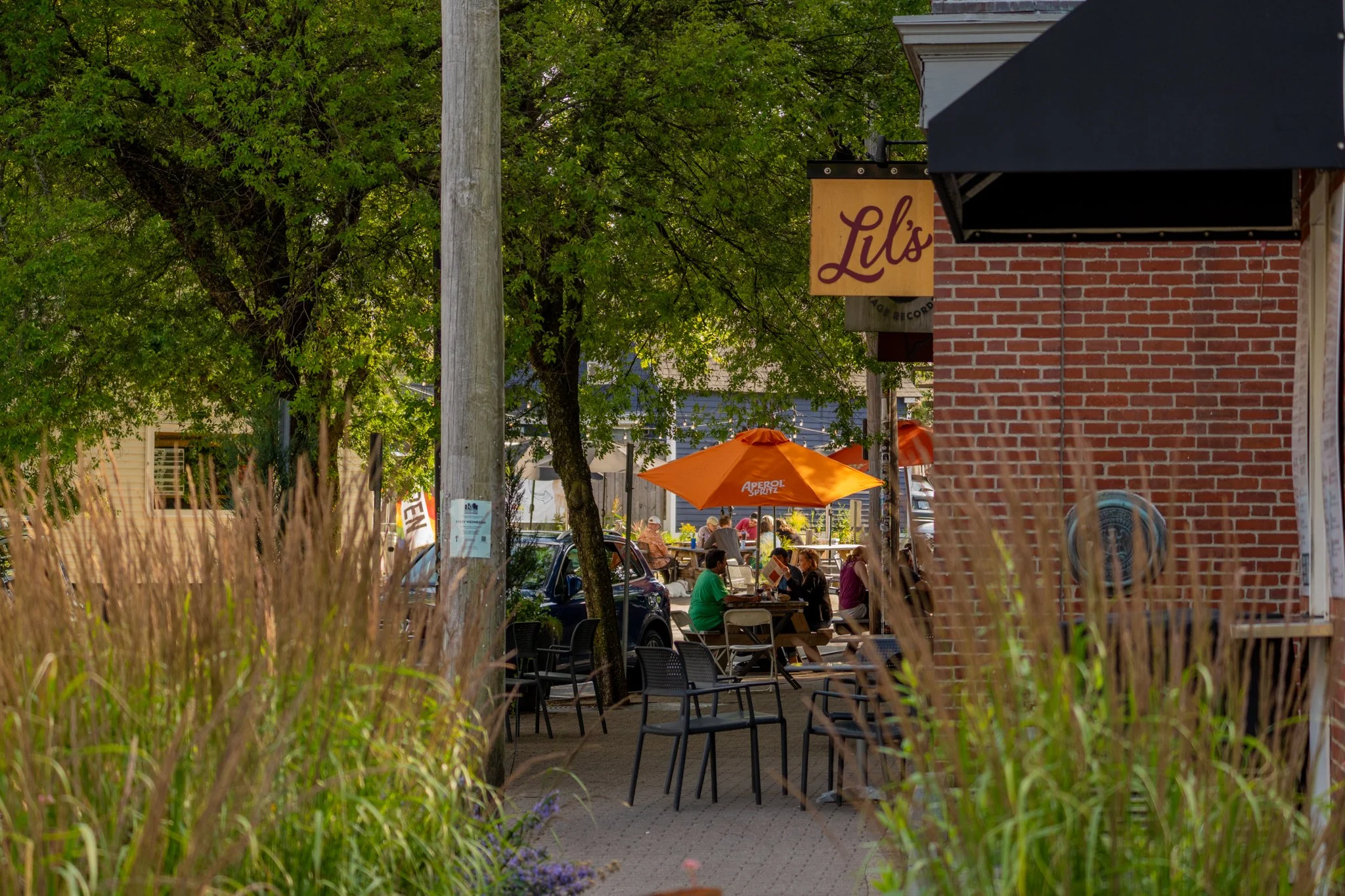 A sidewalk café with outdoor seating under orange umbrellas, people dining, surrounded by green trees, and a brick building with a sign reading "Lil's" in the background.