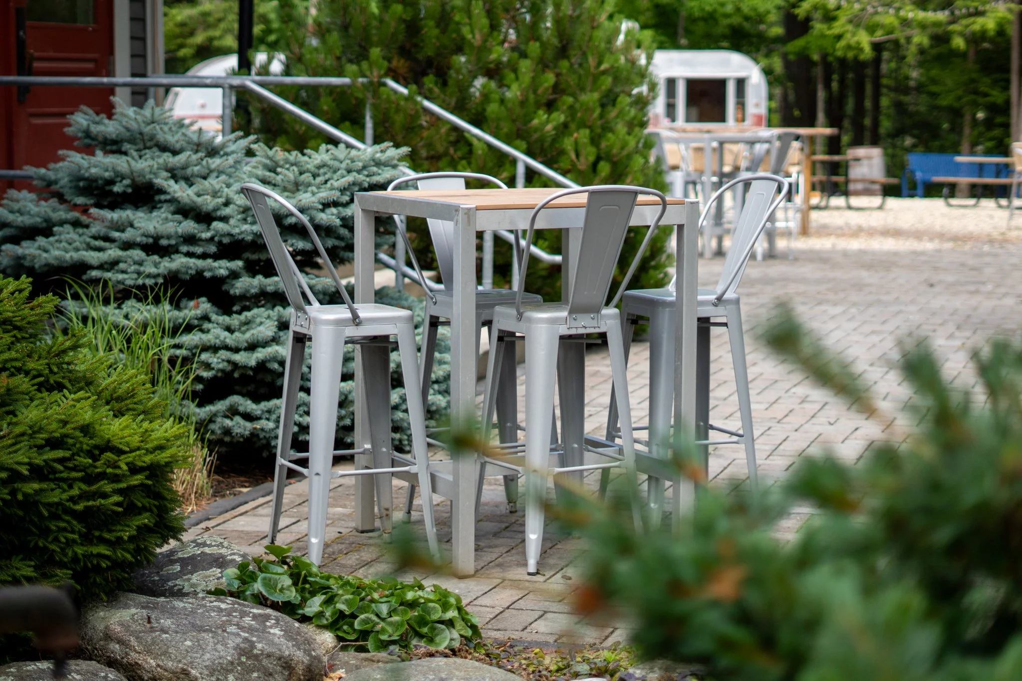 Empty outdoor patio with four metallic chairs and a high-top table, surrounded by green plants, conifer and deciduous trees, with a gravel pathway and additional seating and a camper trailer in the background.