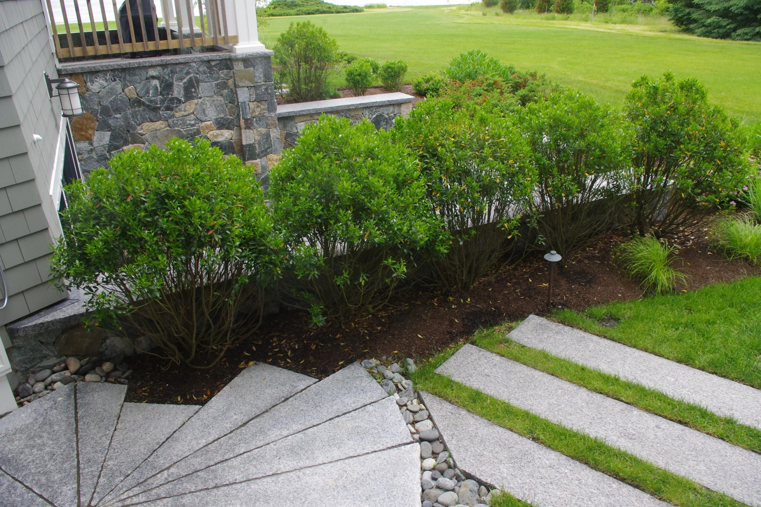 Landscape view of a backyard with a custom stone and gravel stone paving for an outdoor shower, lush green shrubs, grass, and a stone wall next to a house.