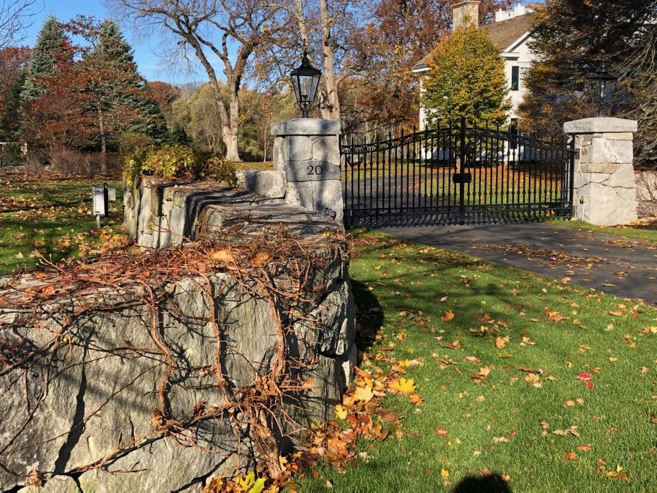 View of a gated estate with stone pillars, a black iron gate, a stone wall, and a white house in the background, surrounded by trees with autumn leaves.