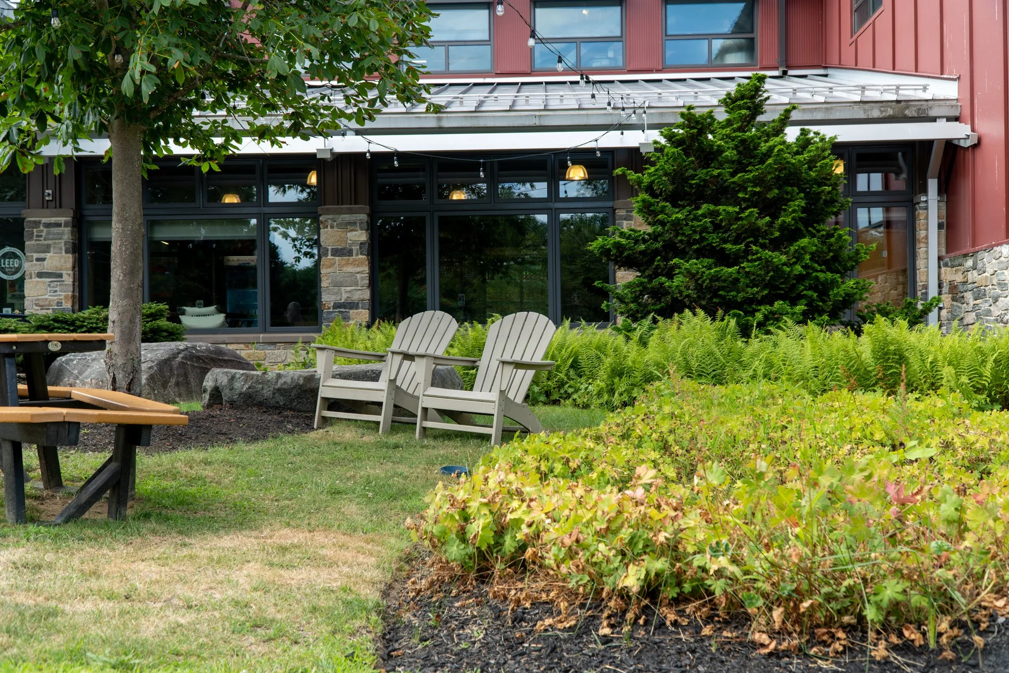 Outdoor cafe patio with two Adirondack chairs facing greenery and a building with large windows and stone accents.