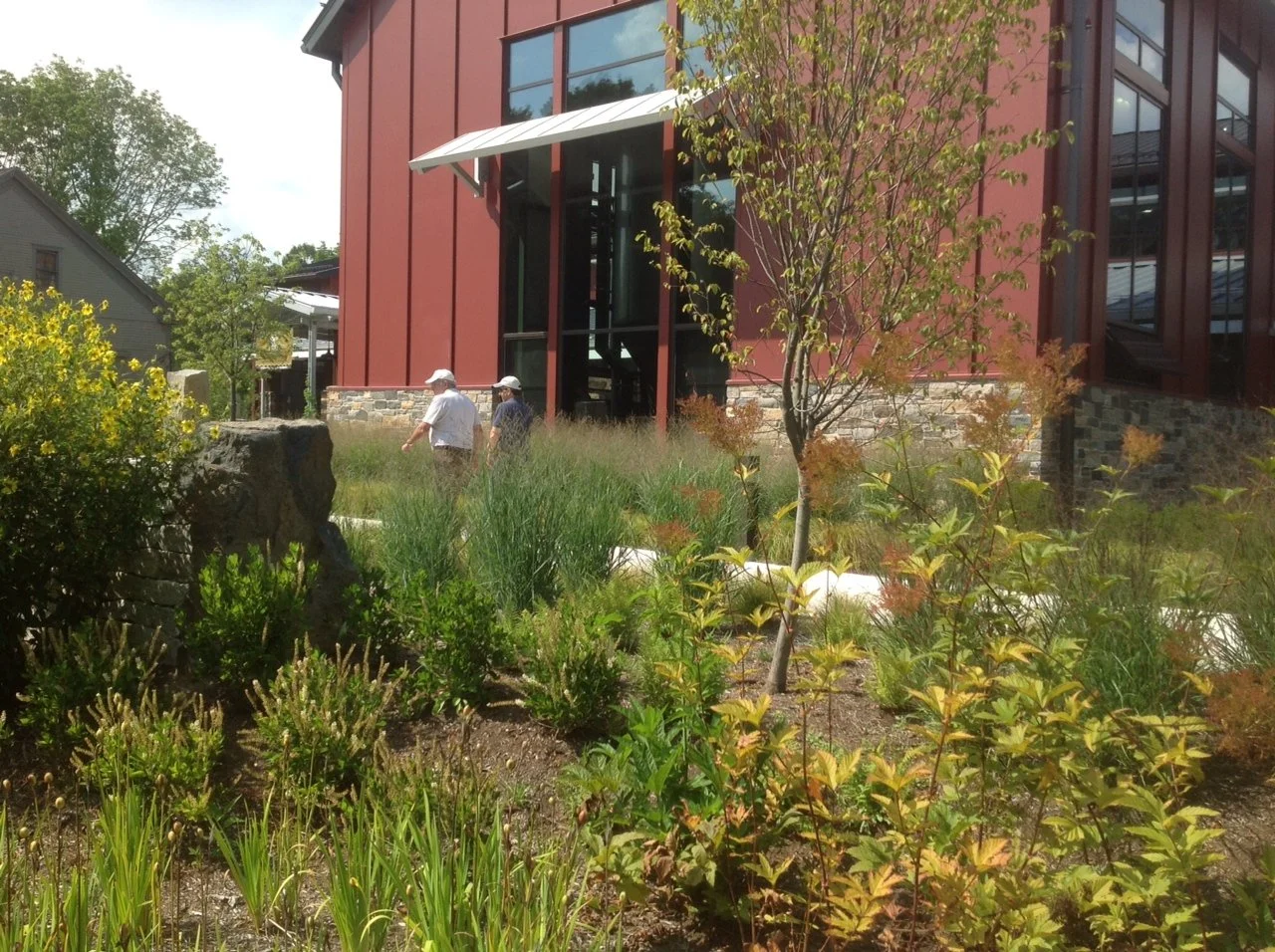 Two men walking through a landscaped garden area in front of a modern building with red exterior walls and large glass windows. The garden features various plants, shrubs, and small trees.