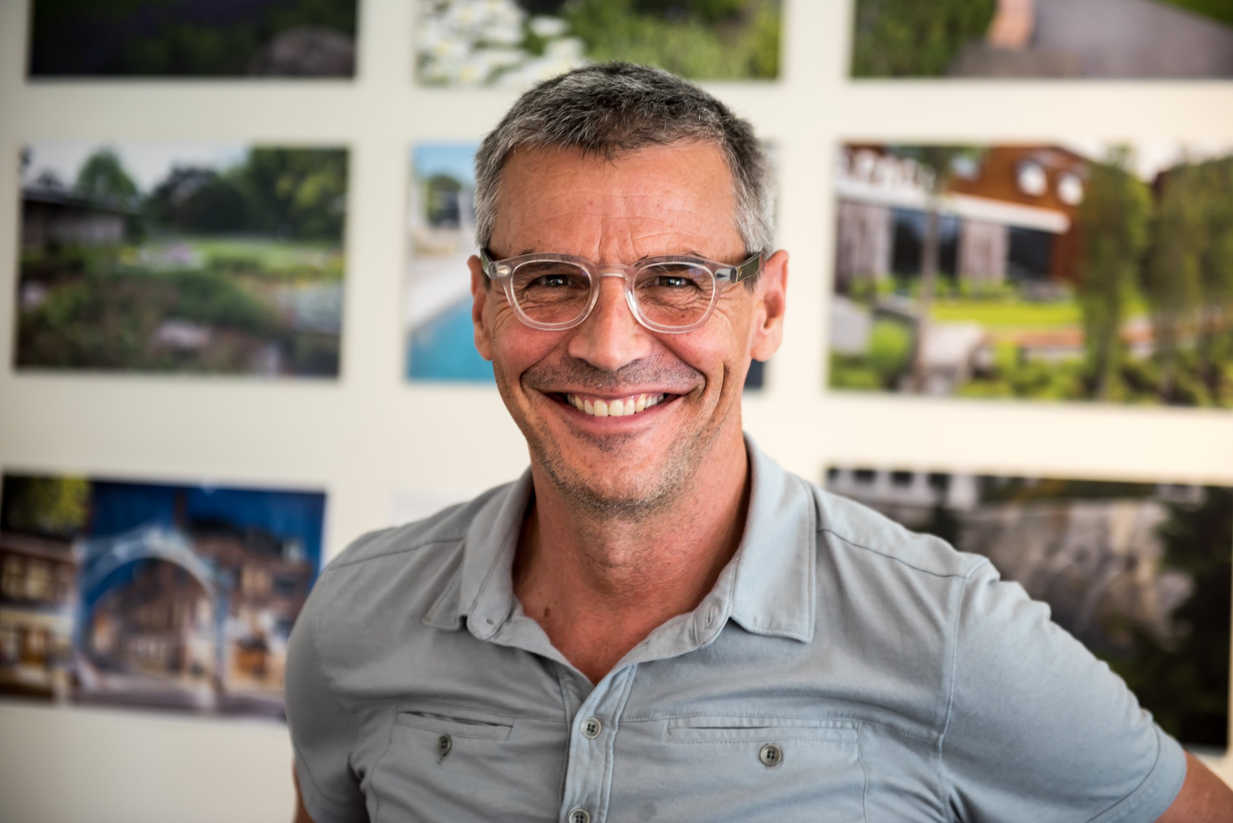 A smiling middle-aged man wearing glasses and a light gray shirt, standing in front of a wall covered with various landscape photographs.