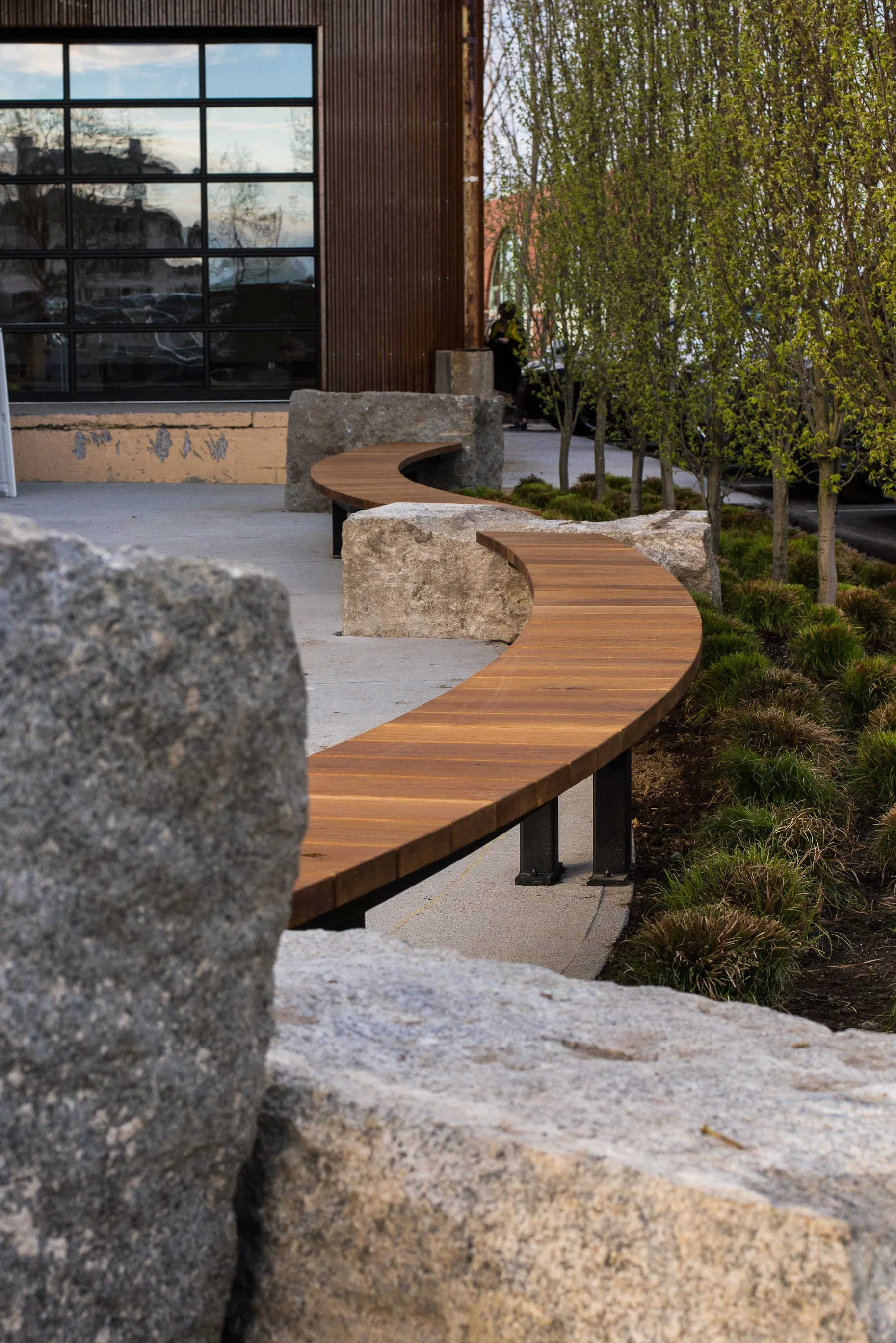 Curved wooden bench supported by black metal legs, situated outdoors among large rocks and small trees, with a building reflecting in the background window.