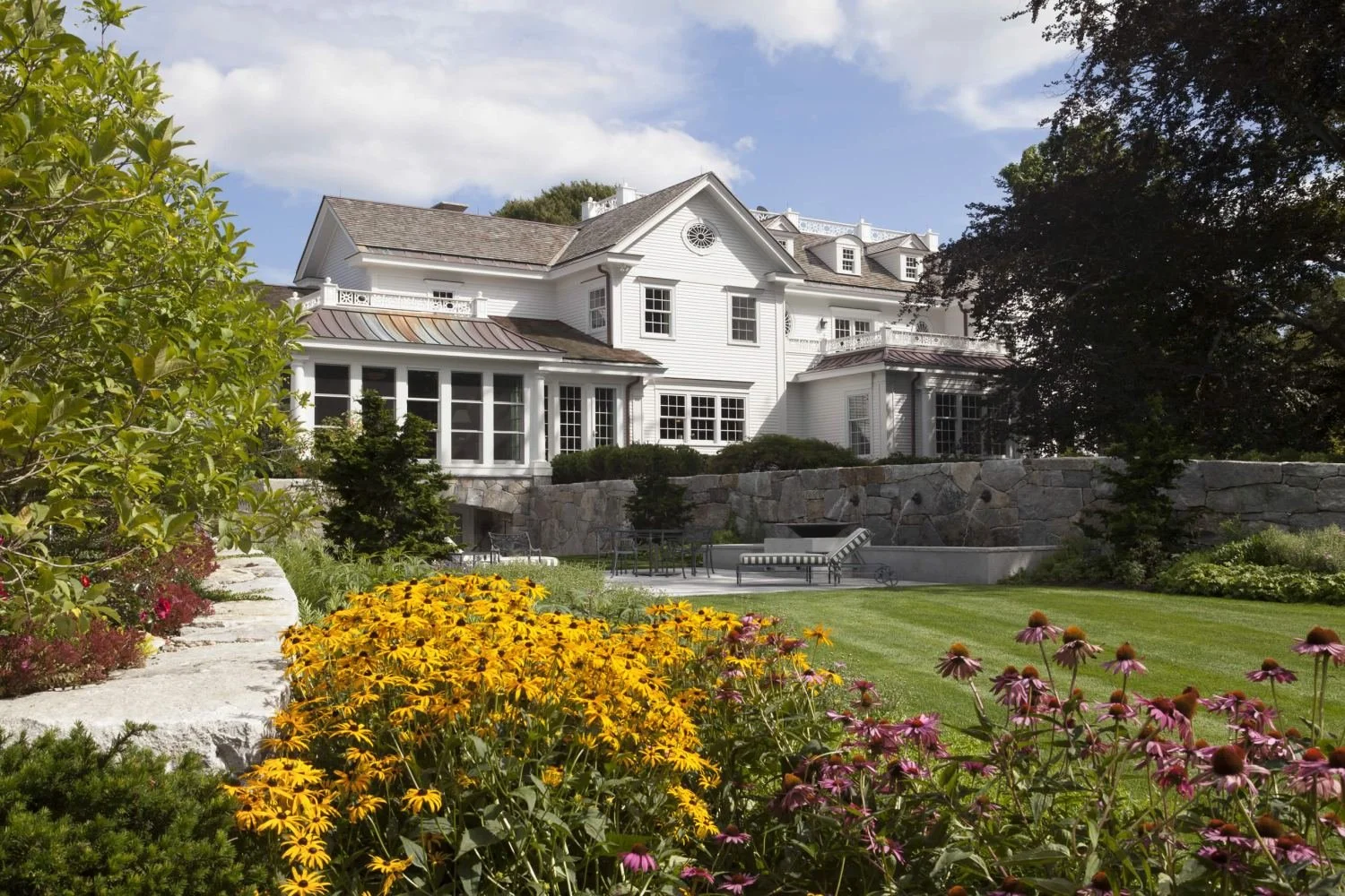A large white house with multiple stories and lots of windows, surrounded by a lush garden with colorful flowers, a green lawn, and trees, under a partly cloudy sky.