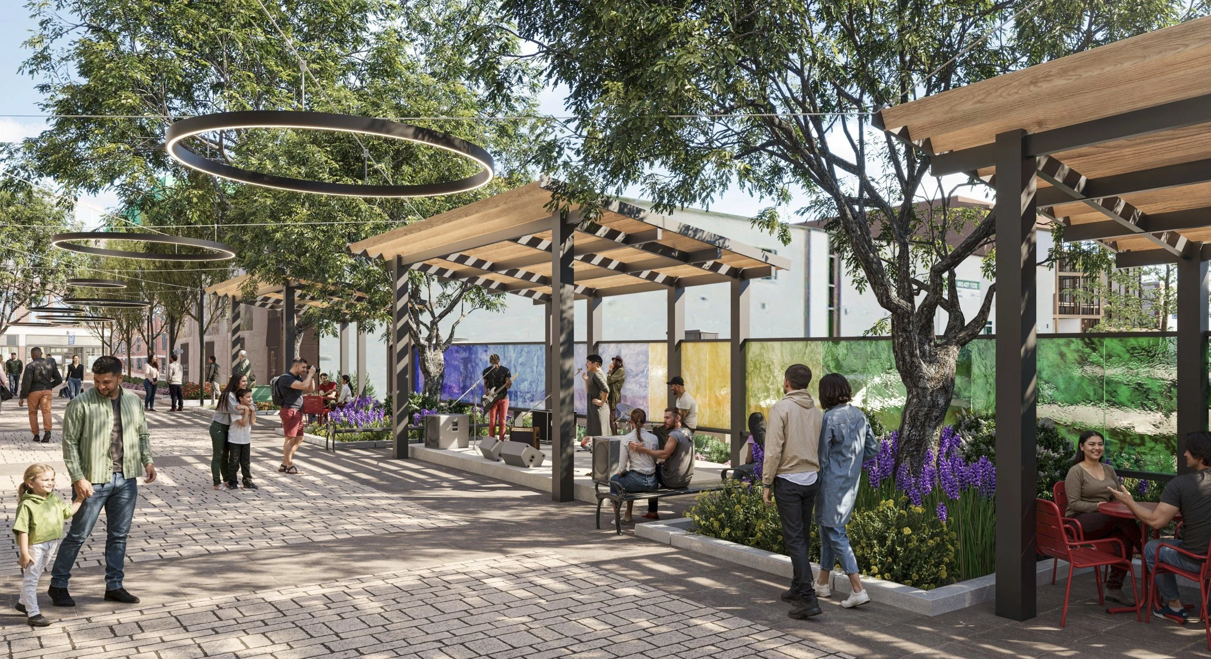 A busy outdoor public space with a small music stage, people sitting, walking, and children playing on a paved walkway surrounded by flowering plants, trees, and modern art installations.