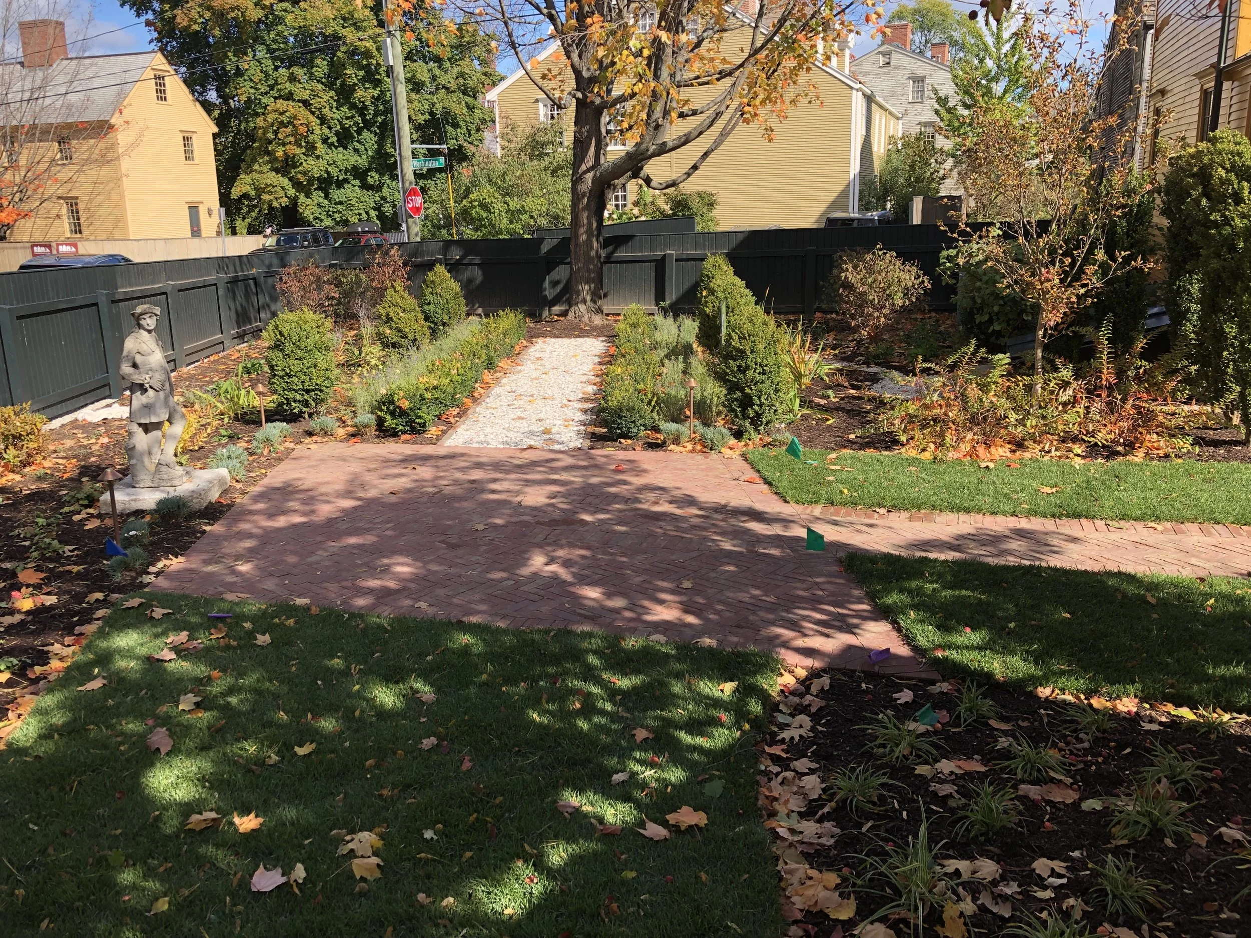 A garden in a residential area with a brick pathway, small green shrubs, a stone statue of a woman, a large tree, and a black fence in the background. Fall leaves are scattered on the ground and the scene is partly shaded by trees.