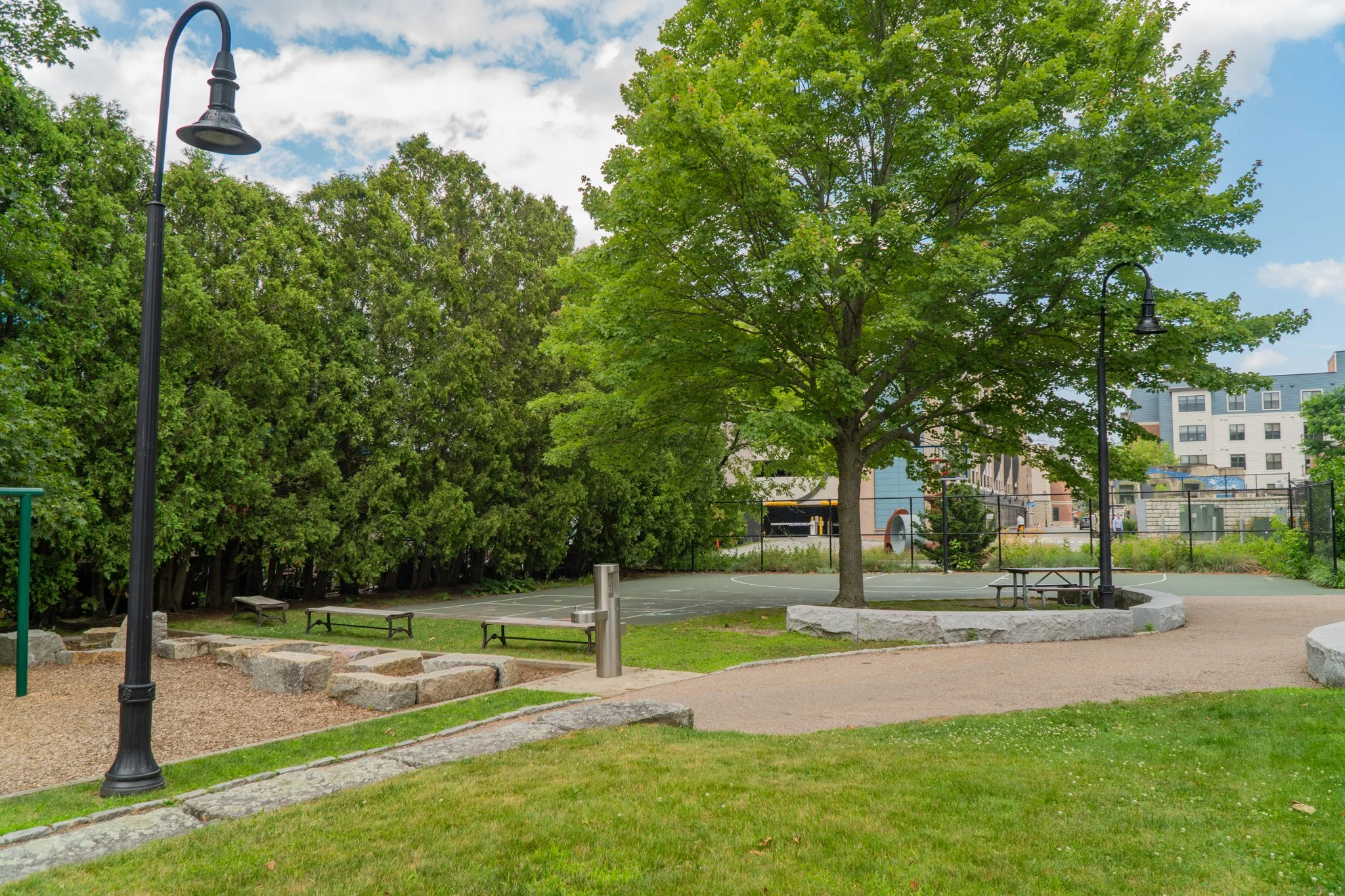 A park with a basketball court, surrounded by green trees and benches, under a partly cloudy sky.
