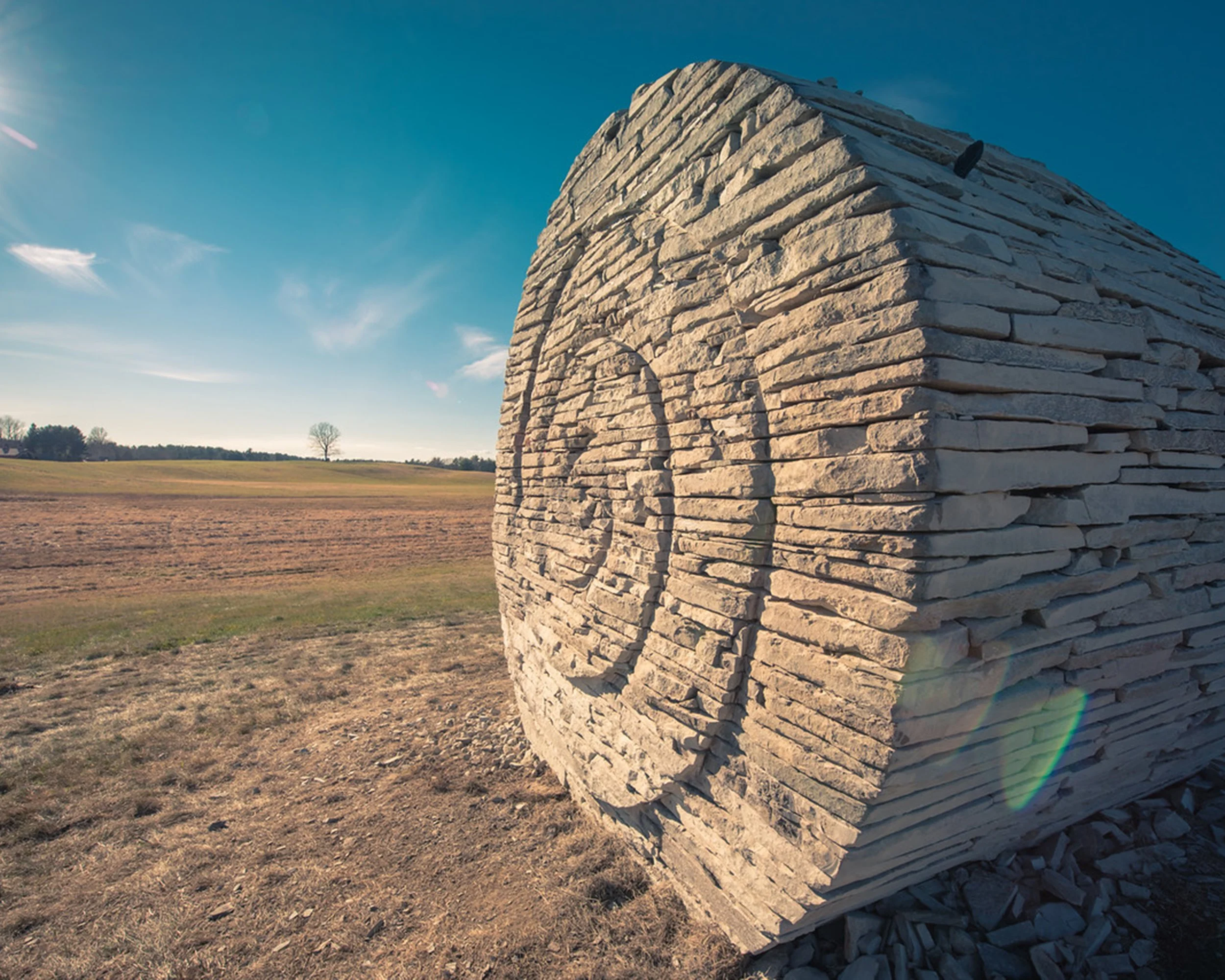 A large stone round haybale sculpture, situated in an open field under a clear blue sky with a few clouds.