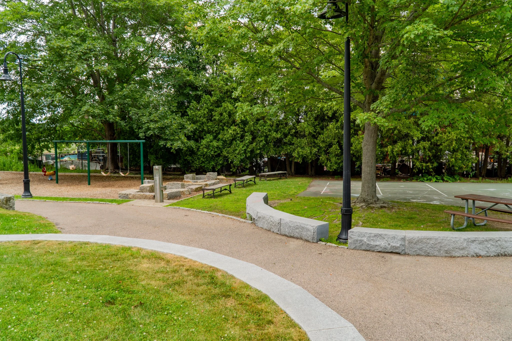 Park with paved walking path, benches, trees, a playground with swings, and a basketball court.