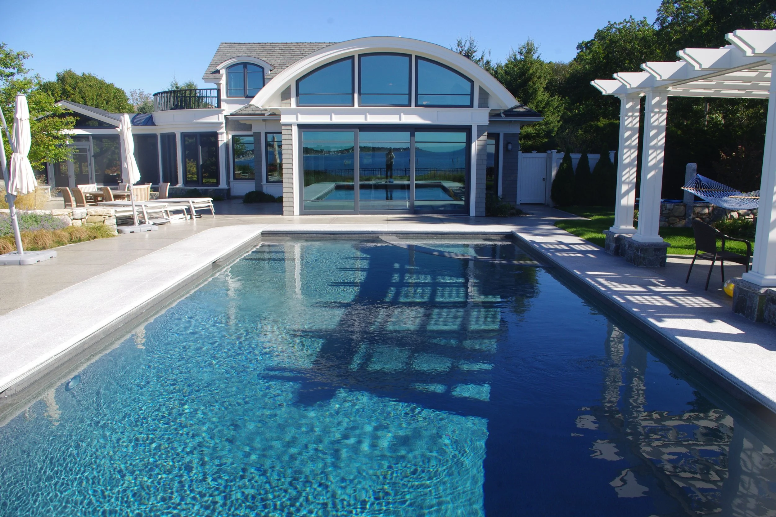 Luxury backyard with a rectangular swimming pool, patio furniture, pergola, and a large modern house with glass doors and windows reflecting the outdoor scene.