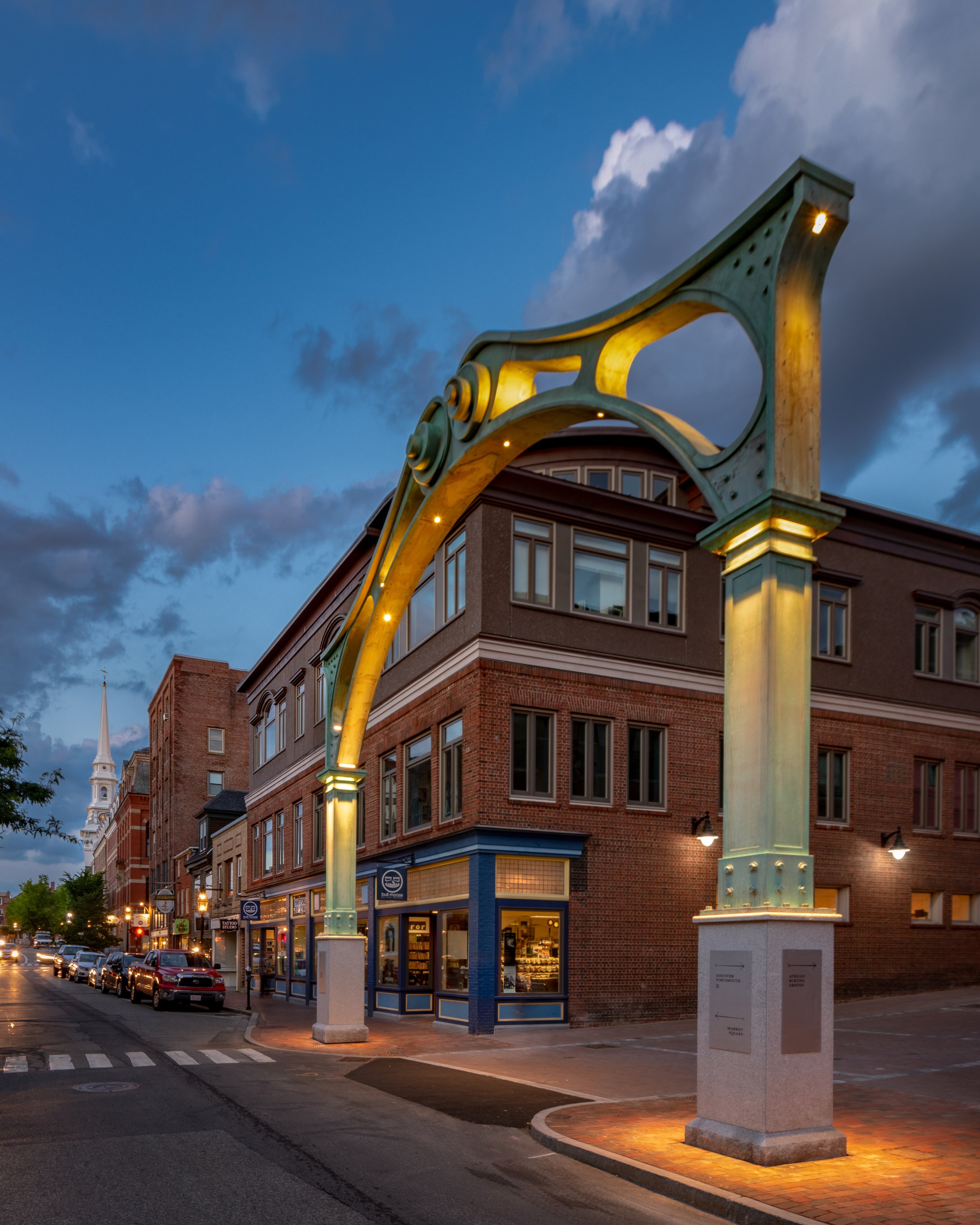 Street scene at dusk with an illuminated green archway on a corner, brick buildings with large windows, parked cars along the street, and a church steeple in the background.