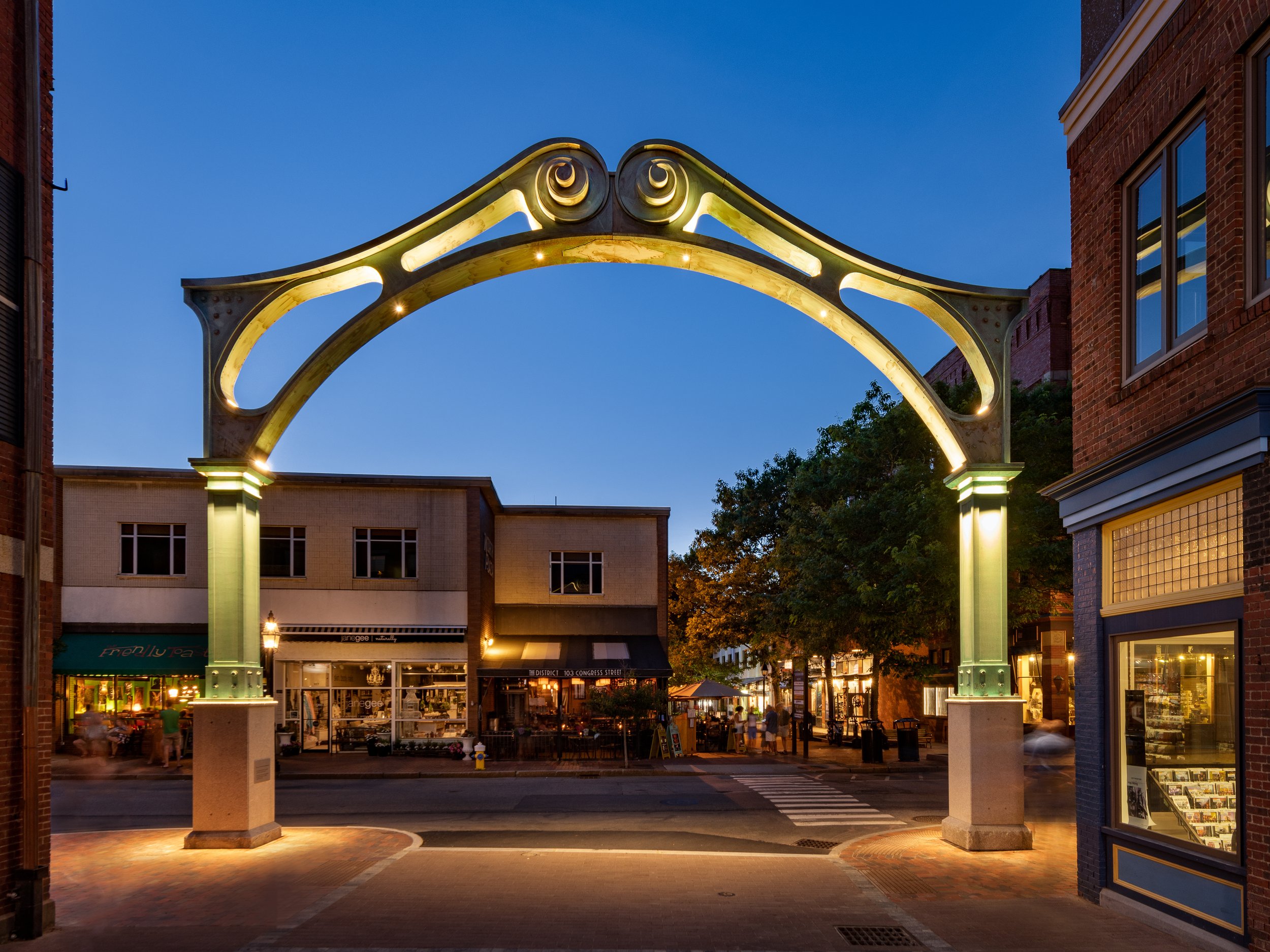 Night view of a city street with an illuminated decorative archway overhead, surrounded by brick and storefront buildings, trees, and people walking.