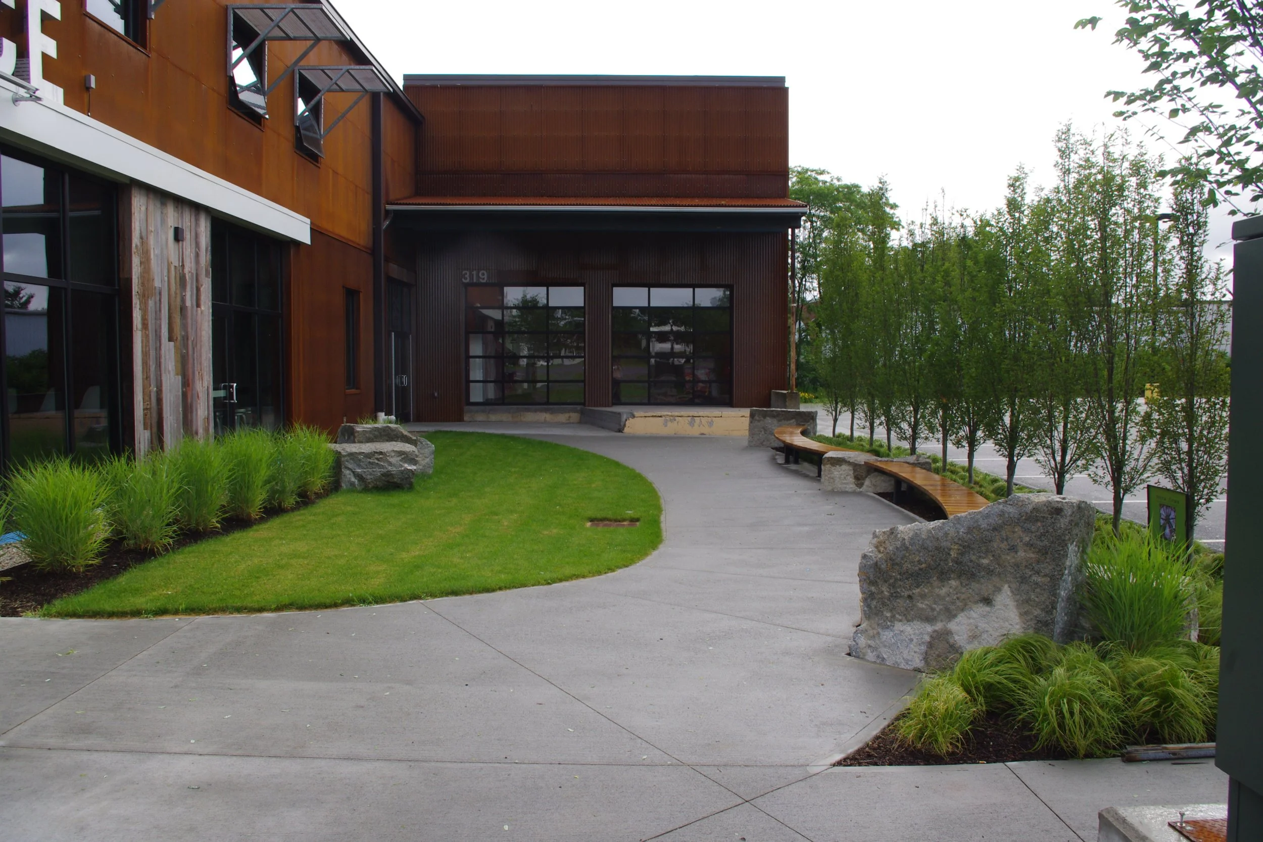 Modern commercial building with a curved sidewalk, green grass, and trees along the edge. Large rocks and benches line the sidewalk near the entrance.
